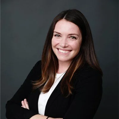 A young woman with dark brown hair, smiling, wearing a black blazer and white top, with arms crossed, against a dark gray background.