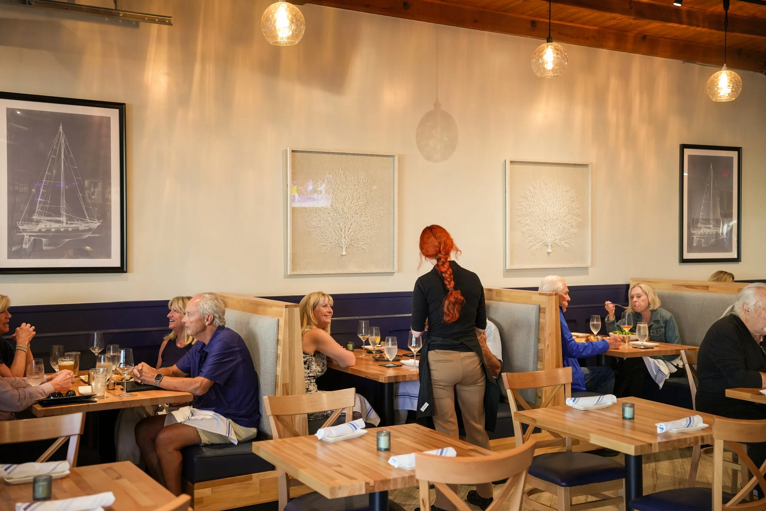People dining in a restaurant with wooden tables, soft candle lighting, and wall art of ships and trees.