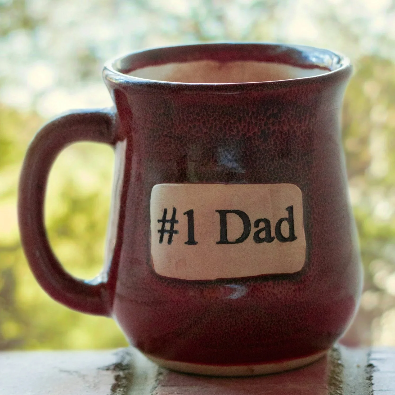 A red ceramic coffee mug with a label that reads '#1 Dad', filled with a hot beverage, placed on a white surface with a blurred outdoor background.