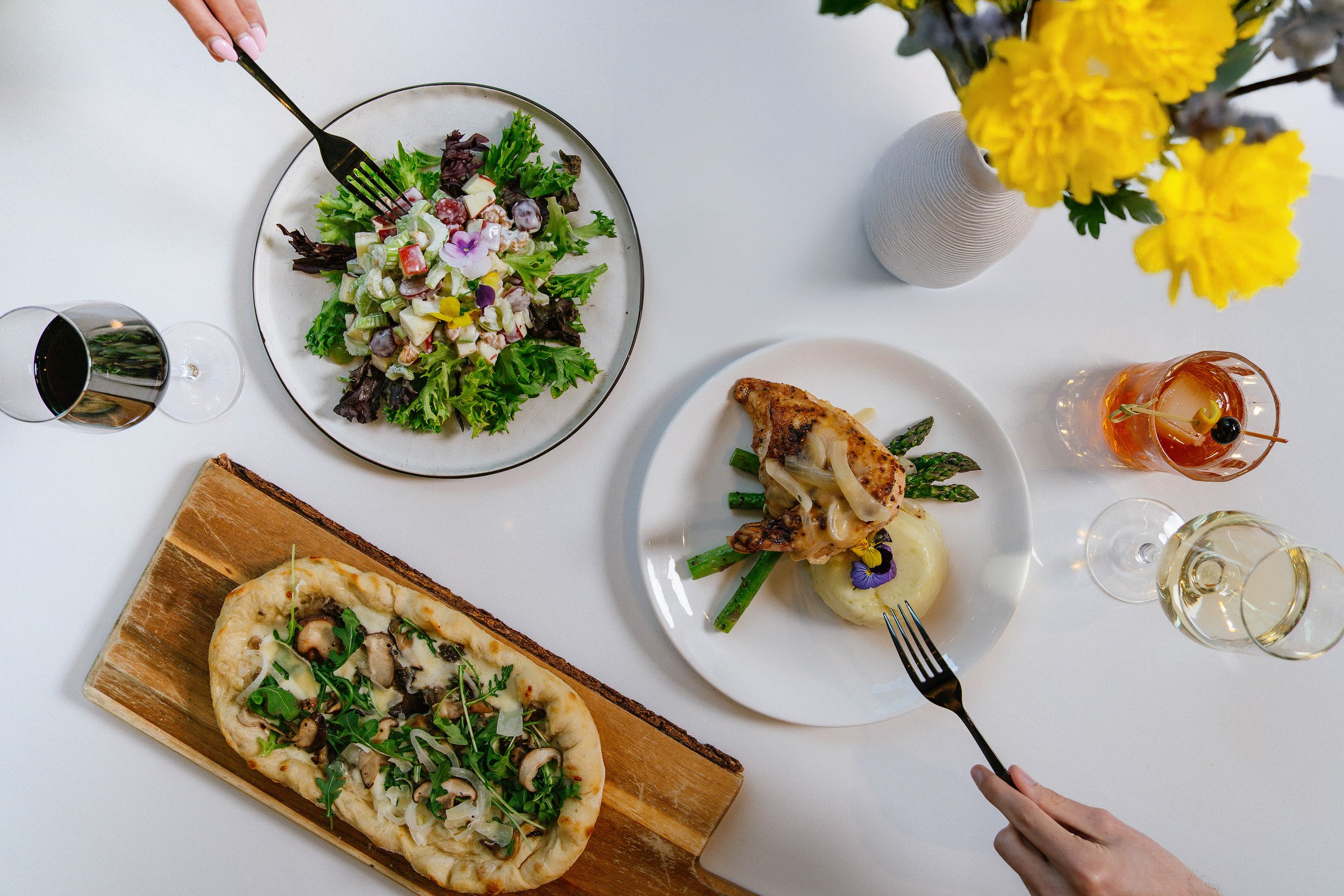 Various plates of food on a white tabletop, including a salad, chicken dish, and pizza.