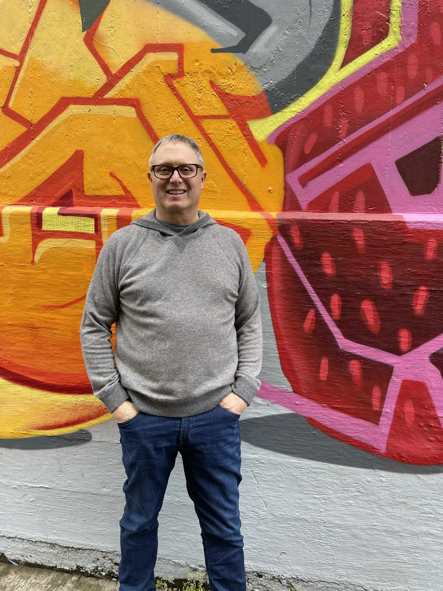 A man standing in front of a colorful graffiti wall, smiling, wearing glasses, a gray hoodie, and blue jeans.