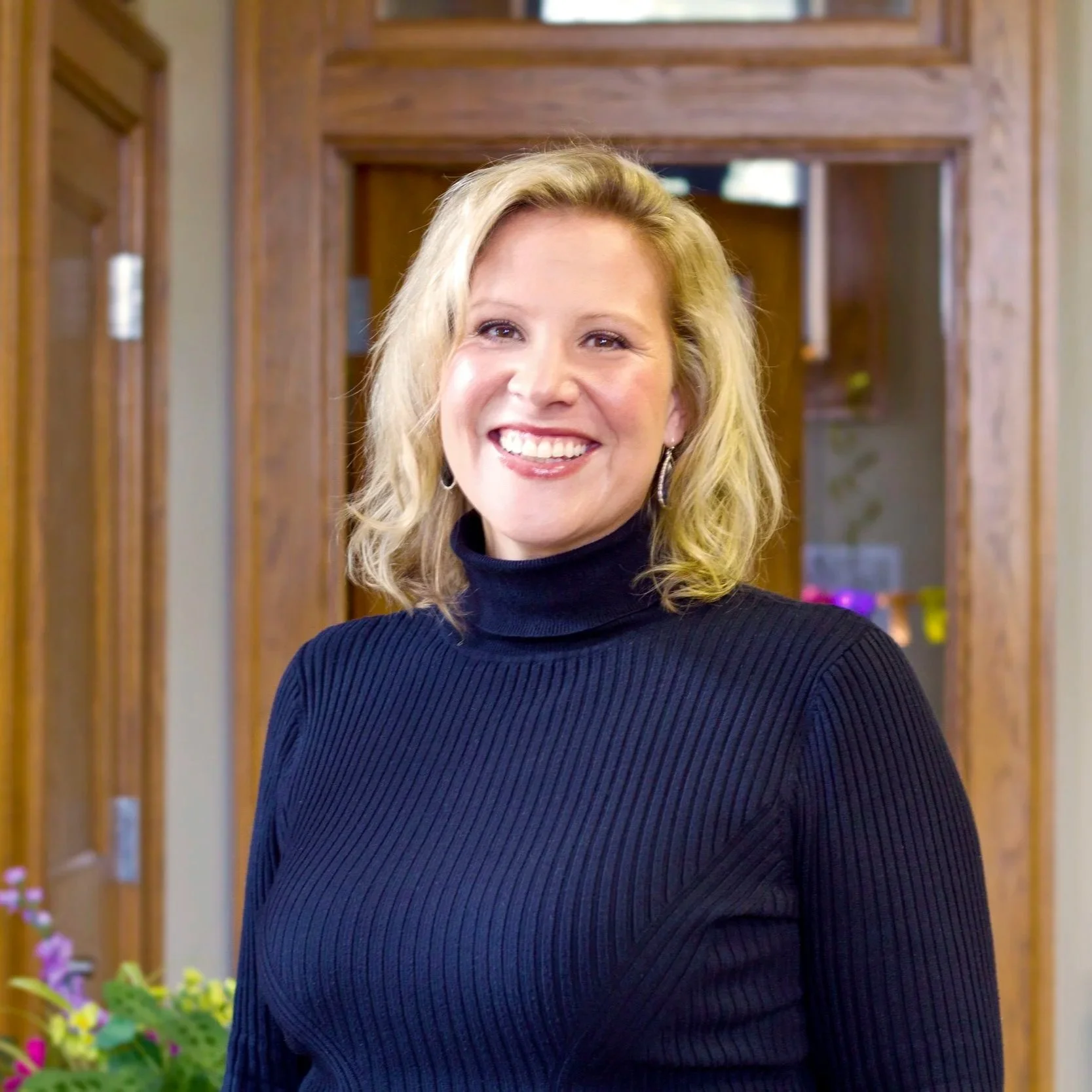 A woman with shoulder-length blonde hair, wearing a black turtleneck sweater, smiling indoors with wooden door frames in the background.