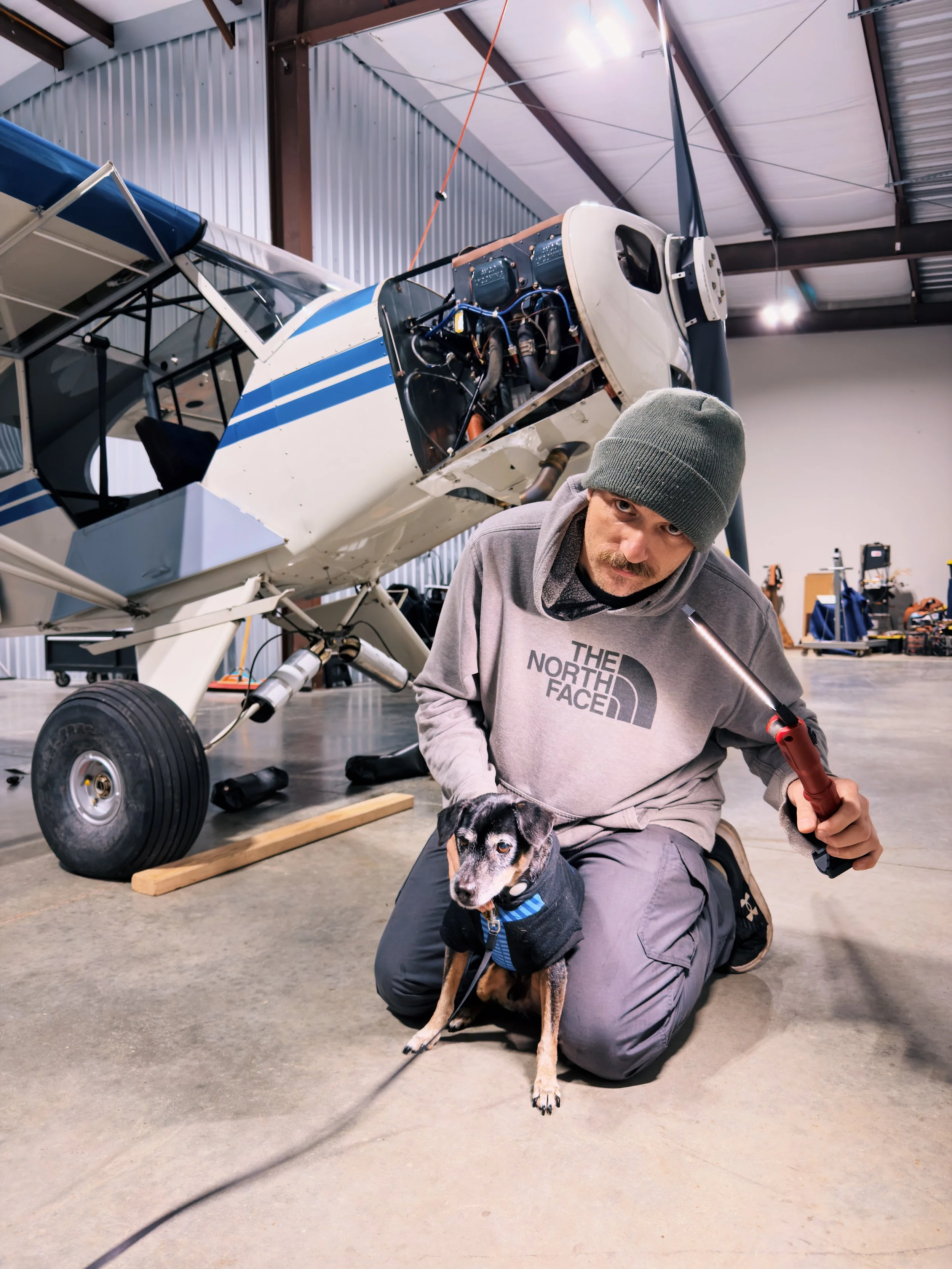 A man wearing sunglasses and a gray shirt working on an aircraft engine outdoors on a sunny day.