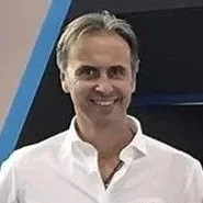 A man with gray hair wearing a white shirt, smiling, standing indoors.