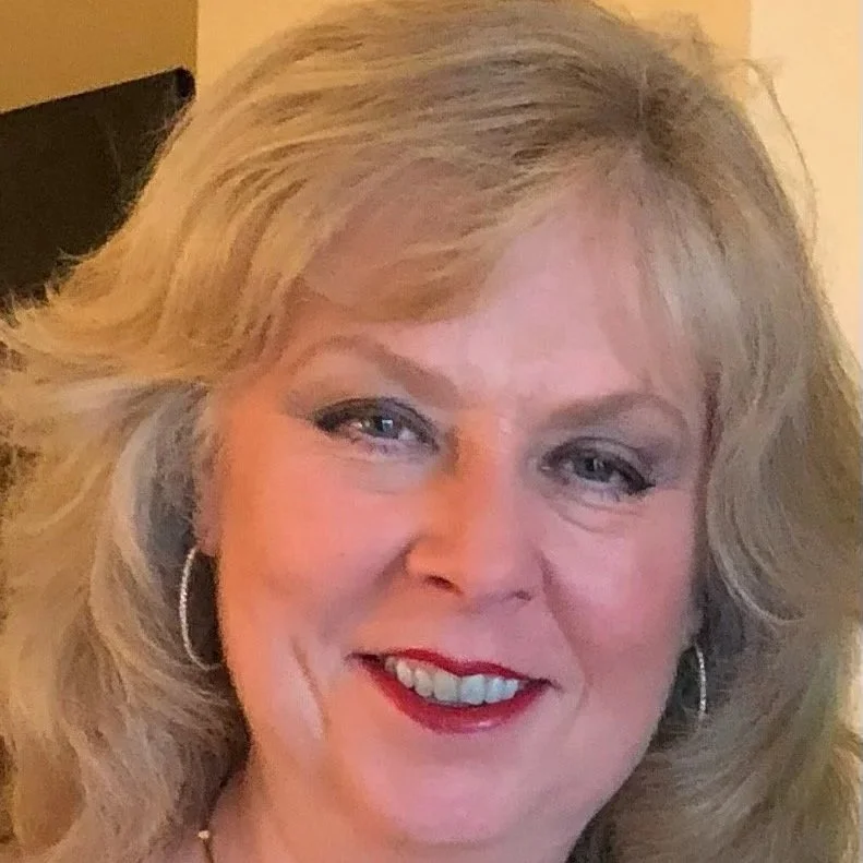 A smiling woman with blonde, wavy hair and hoop earrings, wearing makeup, and standing indoors.