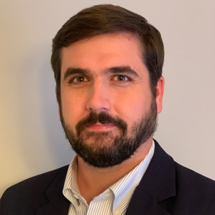 Headshot of a man with dark hair, beard, and mustache, wearing a dark suit jacket and a light striped shirt, against a plain background.