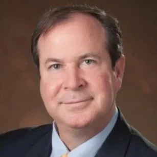 Professional headshot of a middle-aged man in a suit and tie, with a warm brown background.