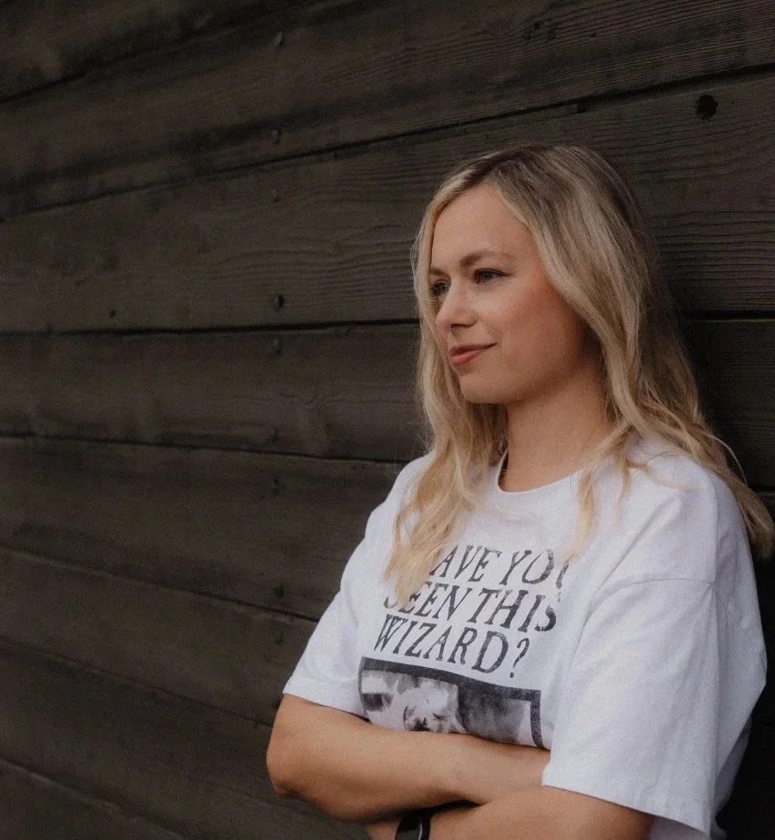 A woman with blonde hair leaning against a dark wooden wall, wearing a white T-shirt that says 'Have you seen this wizard?' with a picture of a cat.