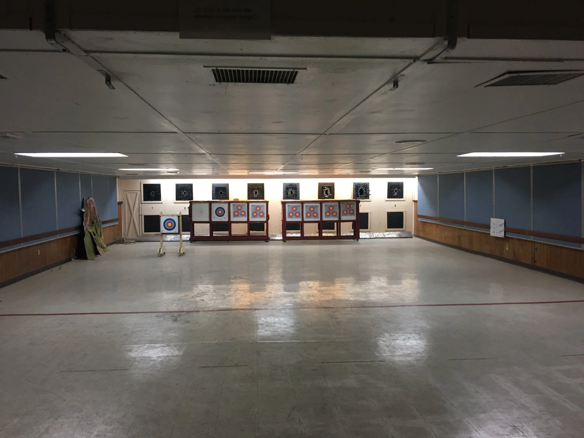 Empty archery shooting range with targets set up at the far end, under fluorescent lighting in an indoor facility.