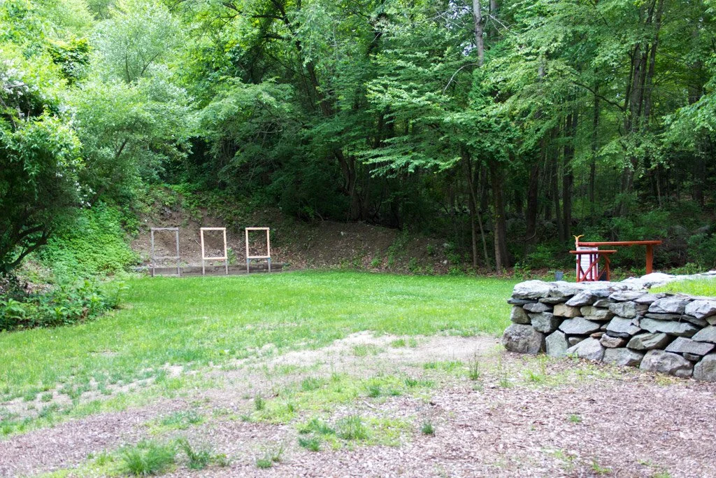 A grassy backyard with a stone retaining wall, a small wooden bridge, and a wooded area in the background.