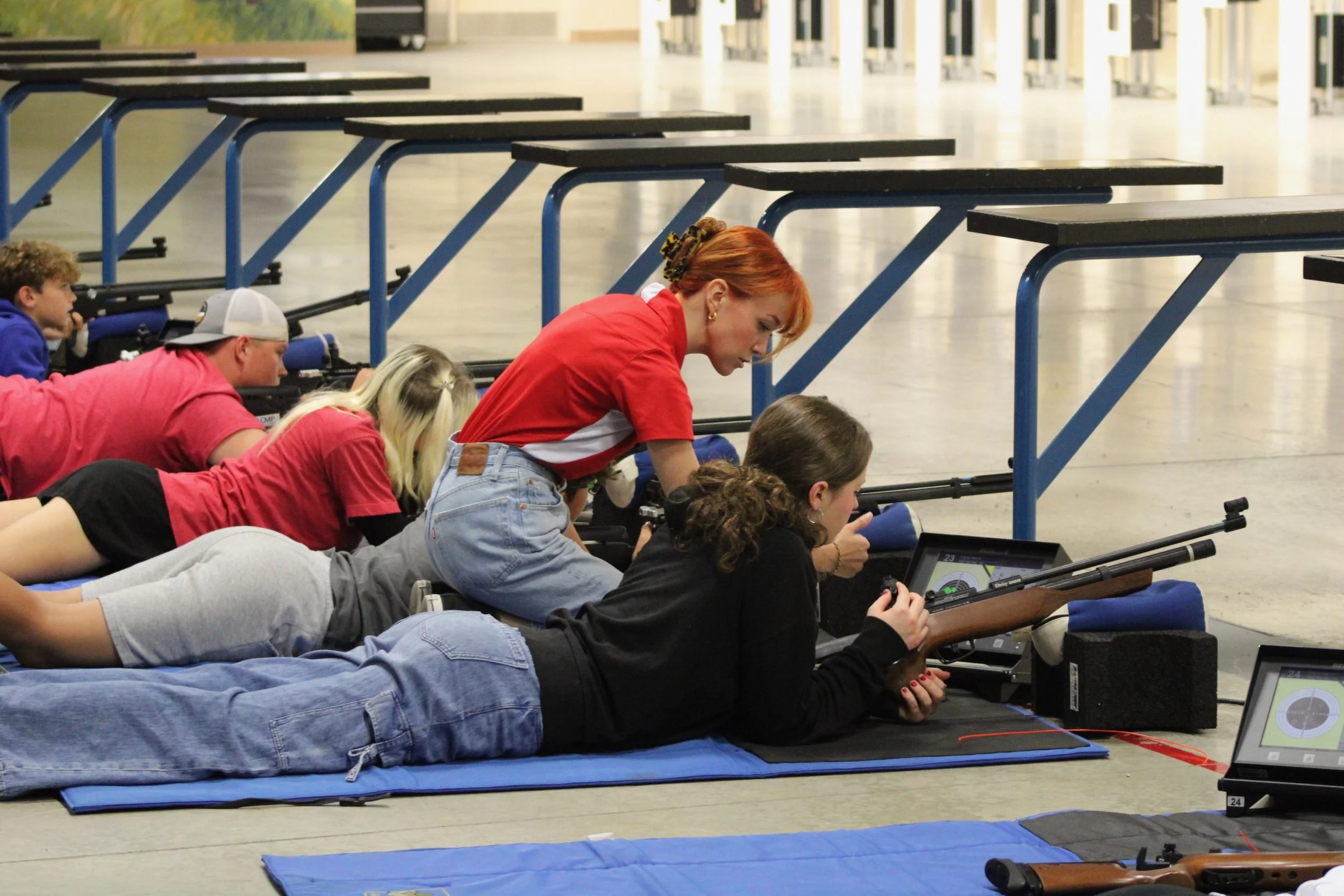 Group of children and a woman lying on the floor, aiming rifles at targets, with electronic targeting devices in front of them, in an indoor shooting range.