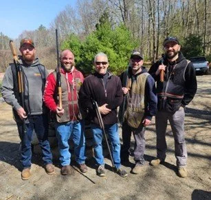 Five men standing outdoors on a dirt path, dressed in casual outdoor clothing, holding walking sticks, with trees and parked cars in the background.