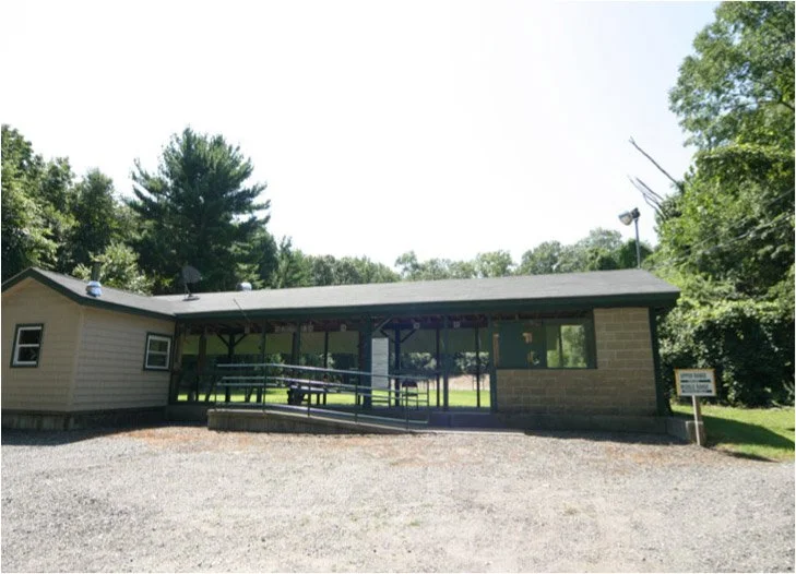 Single-story building with large glass windows and a sloped roof, surrounded by green trees and a gravel parking area.