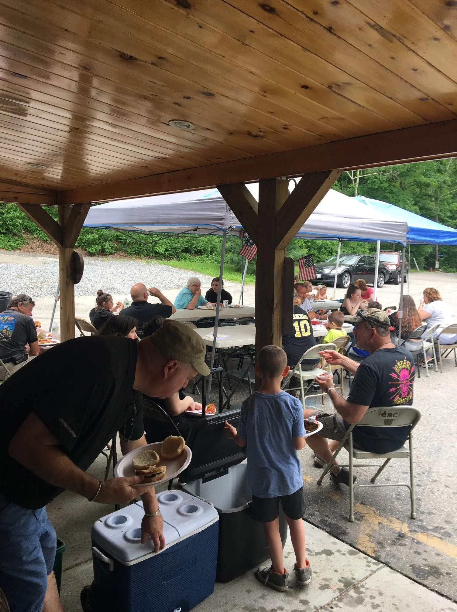 People gathered under a wooden pavilion, sitting at tables, eating and socializing. Adults and children are present, with some wearing casual clothes and baseball caps. A man is serving food from a cooler, and there are tents and vehicles in the background.