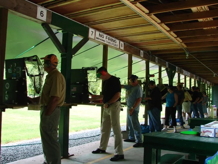 People standing in line at a shooting range, aiming guns at targets, outdoor covered shooting lanes with numbered signs and a sign that reads 'No Firing Beyond This Point'.