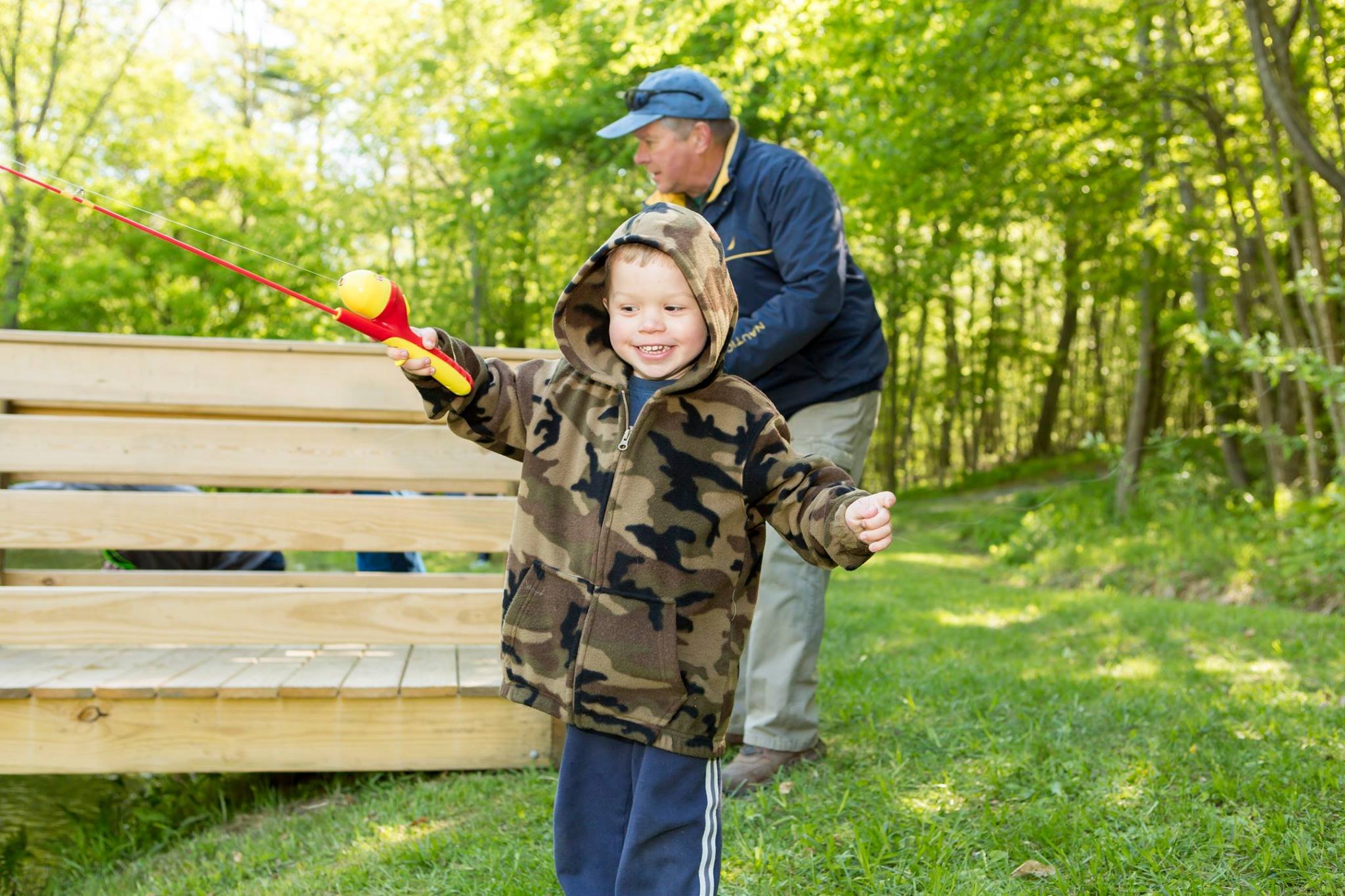 A young boy in a camouflage hoodie holding a toy fishing rod with a yellow and red ball on the end, standing outdoors in front of a wooden structure and a man fishing in a green, wooded area.
