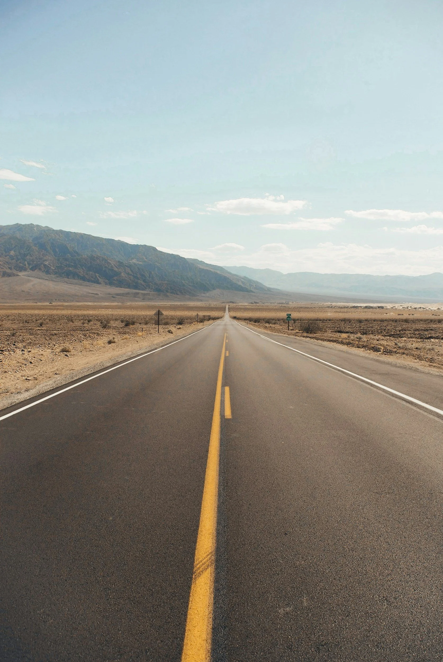 Open desert highway extending straight into the horizon with mountains in the distance, under a partly cloudy sky.