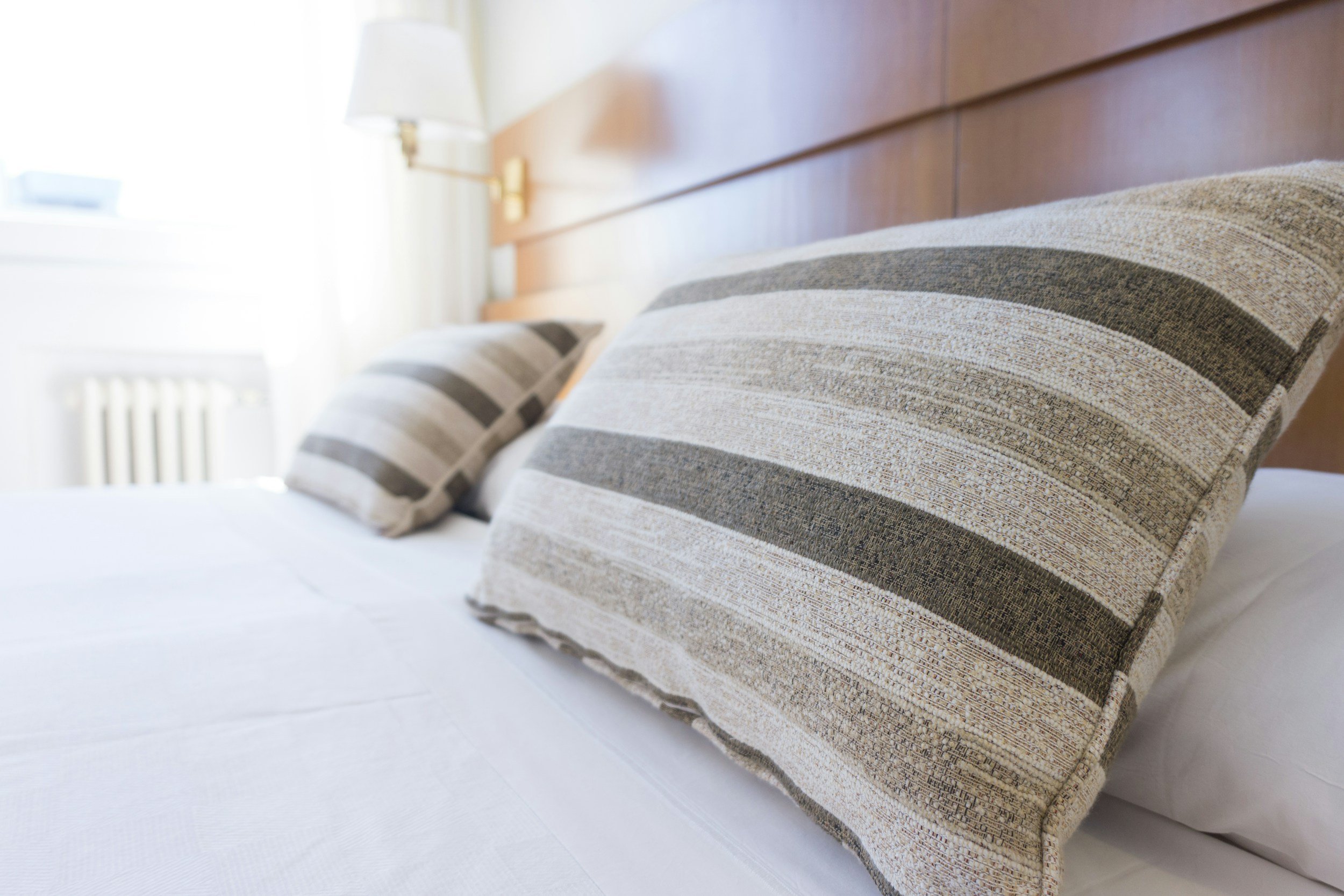 Close-up of a bed with two striped pillows resting against a wooden headboard in a brightly lit hotel room.