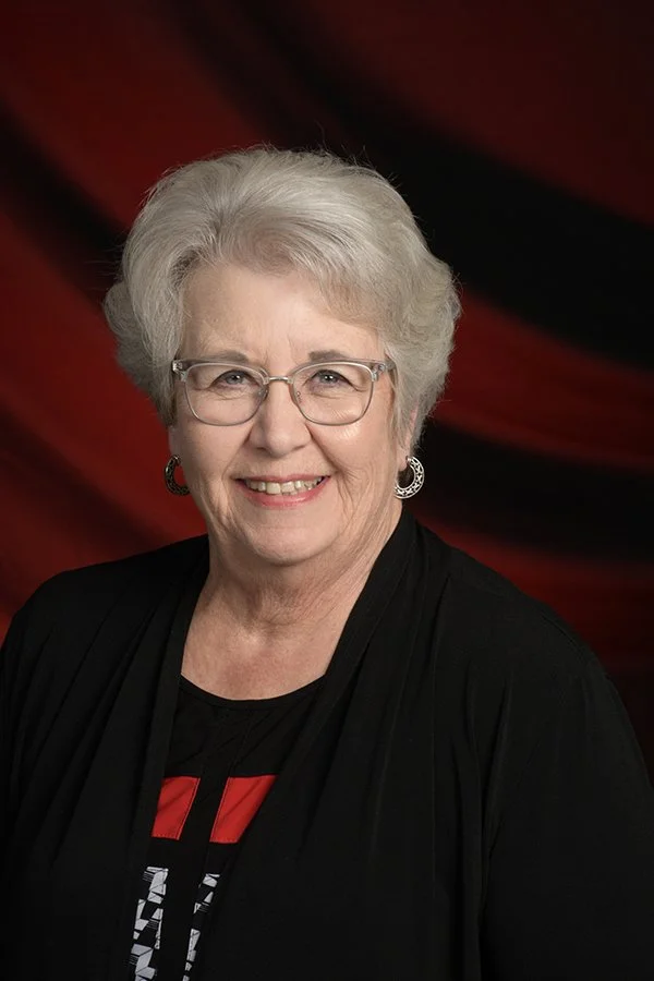 A portrait of an elderly woman with short white hair, glasses, and earrings, smiling at the camera, wearing a black top with red and white details, against a dark red and black background.
