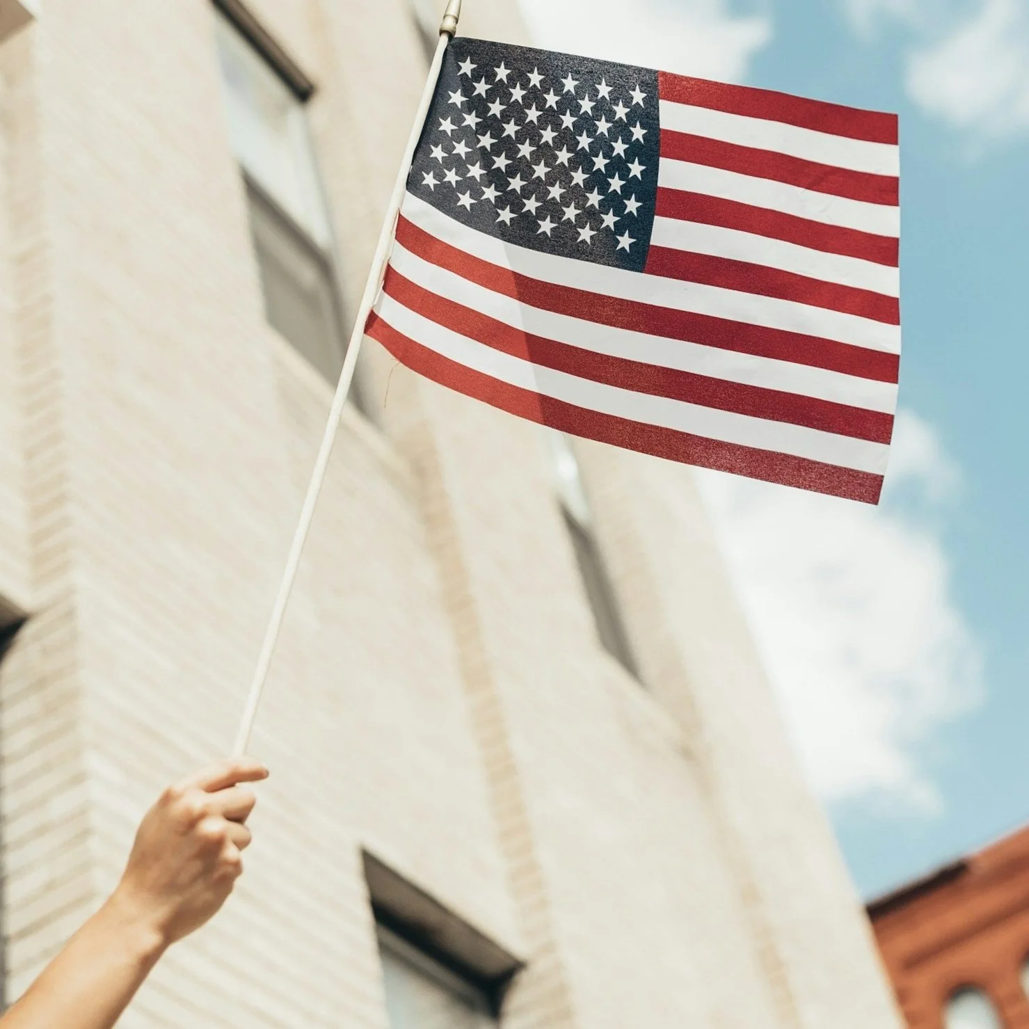 Person holding an American flag on a pole outside a building with a beige brick wall and windows, under a partly cloudy sky.