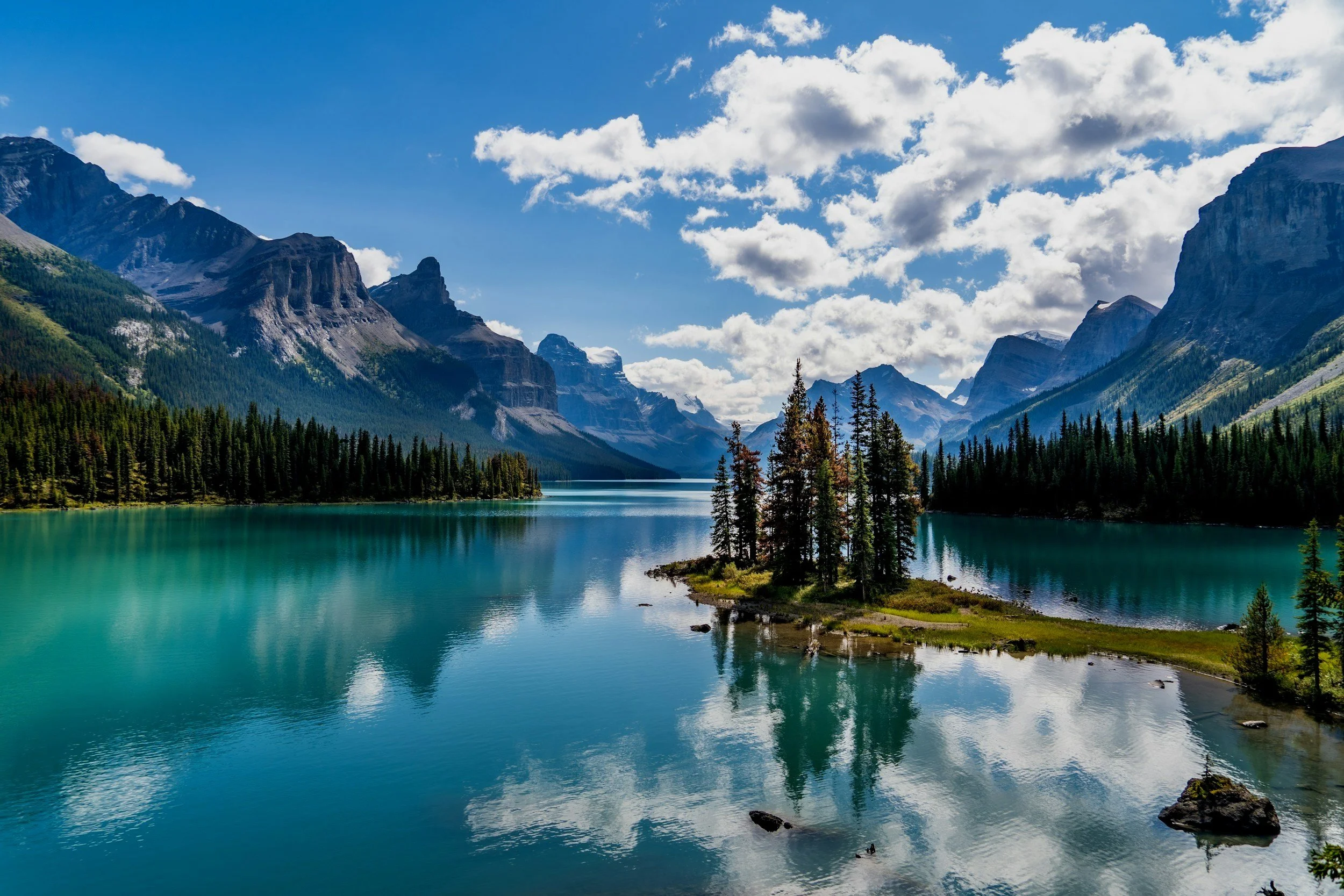 A scenic view of a turquoise lake surrounded by lush green forests and tall mountains under a partly cloudy sky.