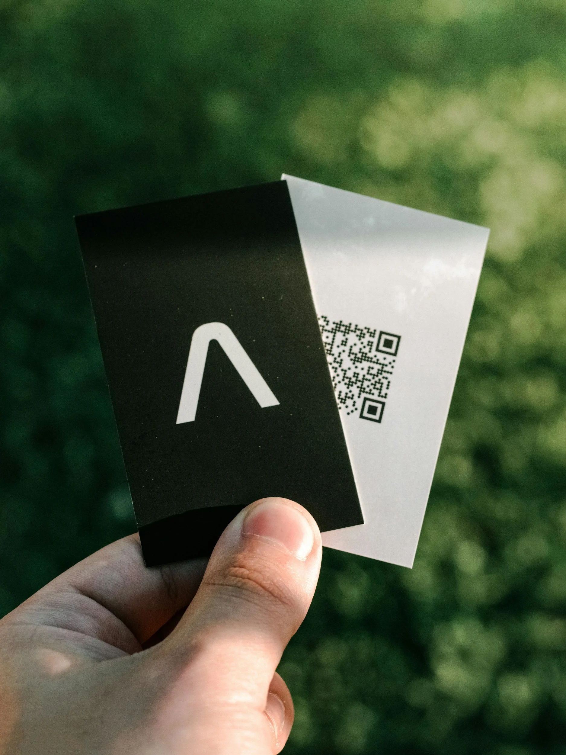 A person's hand holding a black and white card with a logo and QR code, with a blurred green outdoor background.