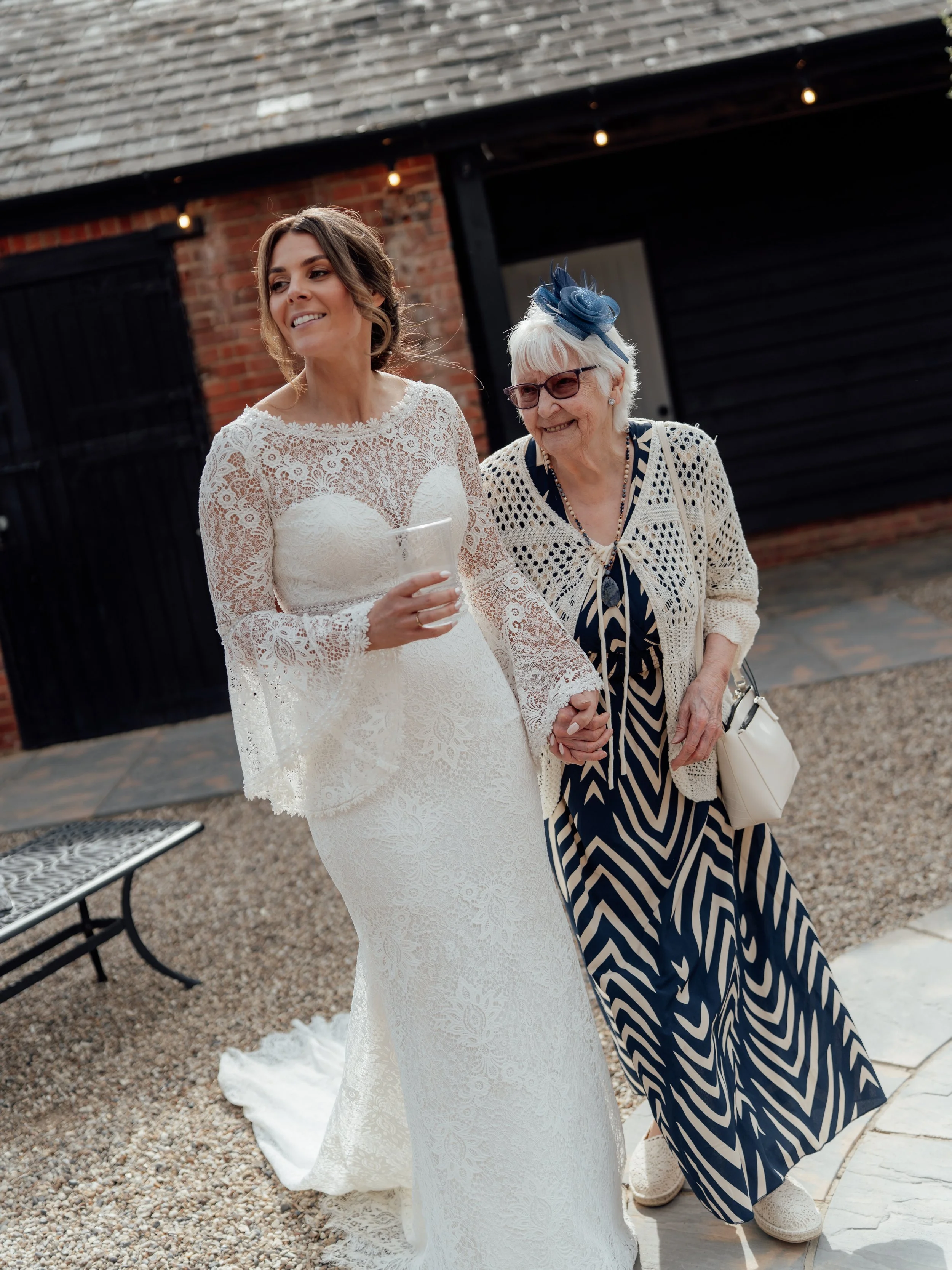 A bride in a white lace wedding dress holding hands and walking with an elderly woman wearing sunglasses and a blue patterned dress with a matching hat, outside near a brick building.