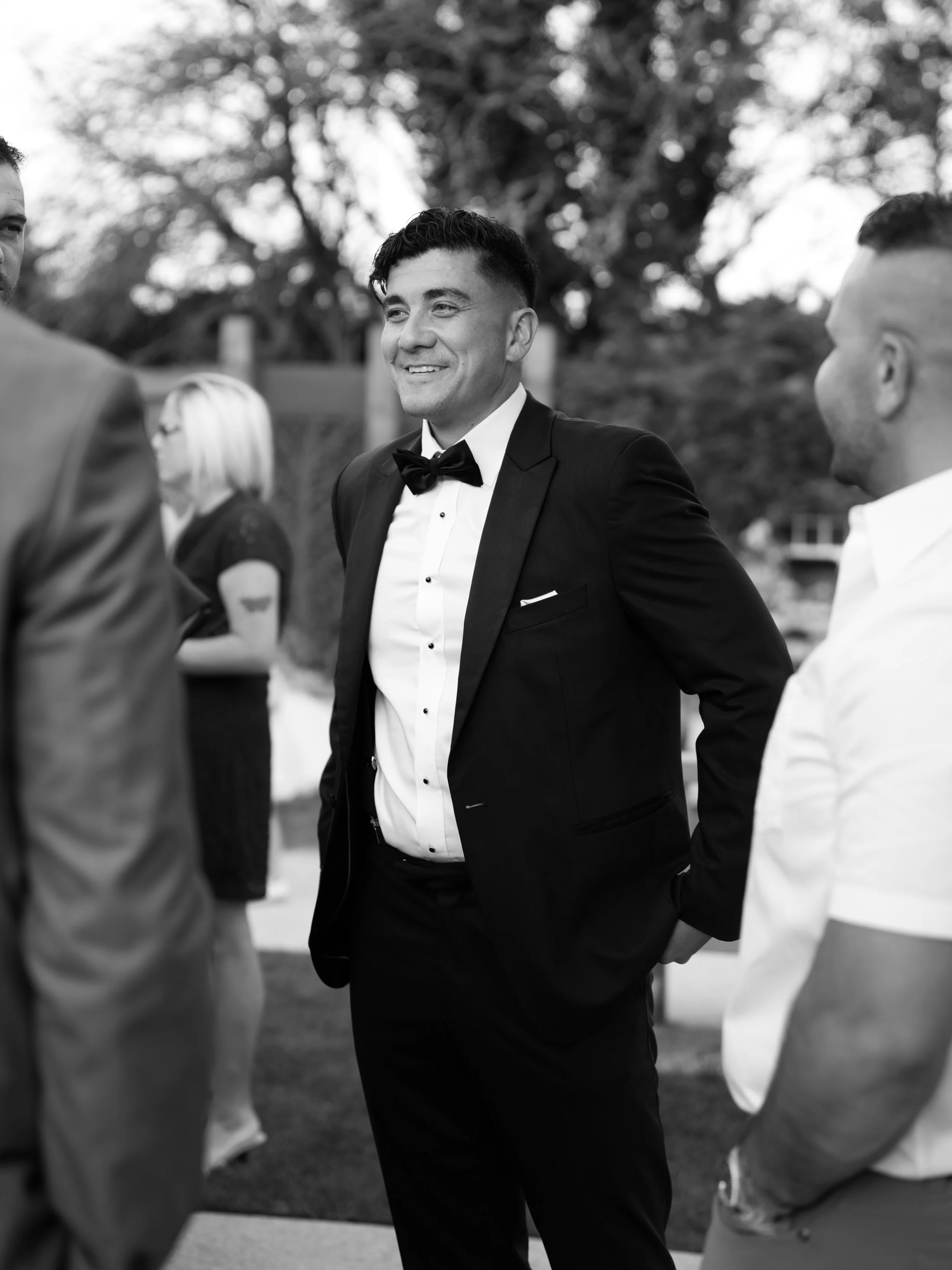 A man in a black tuxedo and bow tie is smiling and talking to a group of people outdoors. The image is in black and white, with trees and other people in the background.