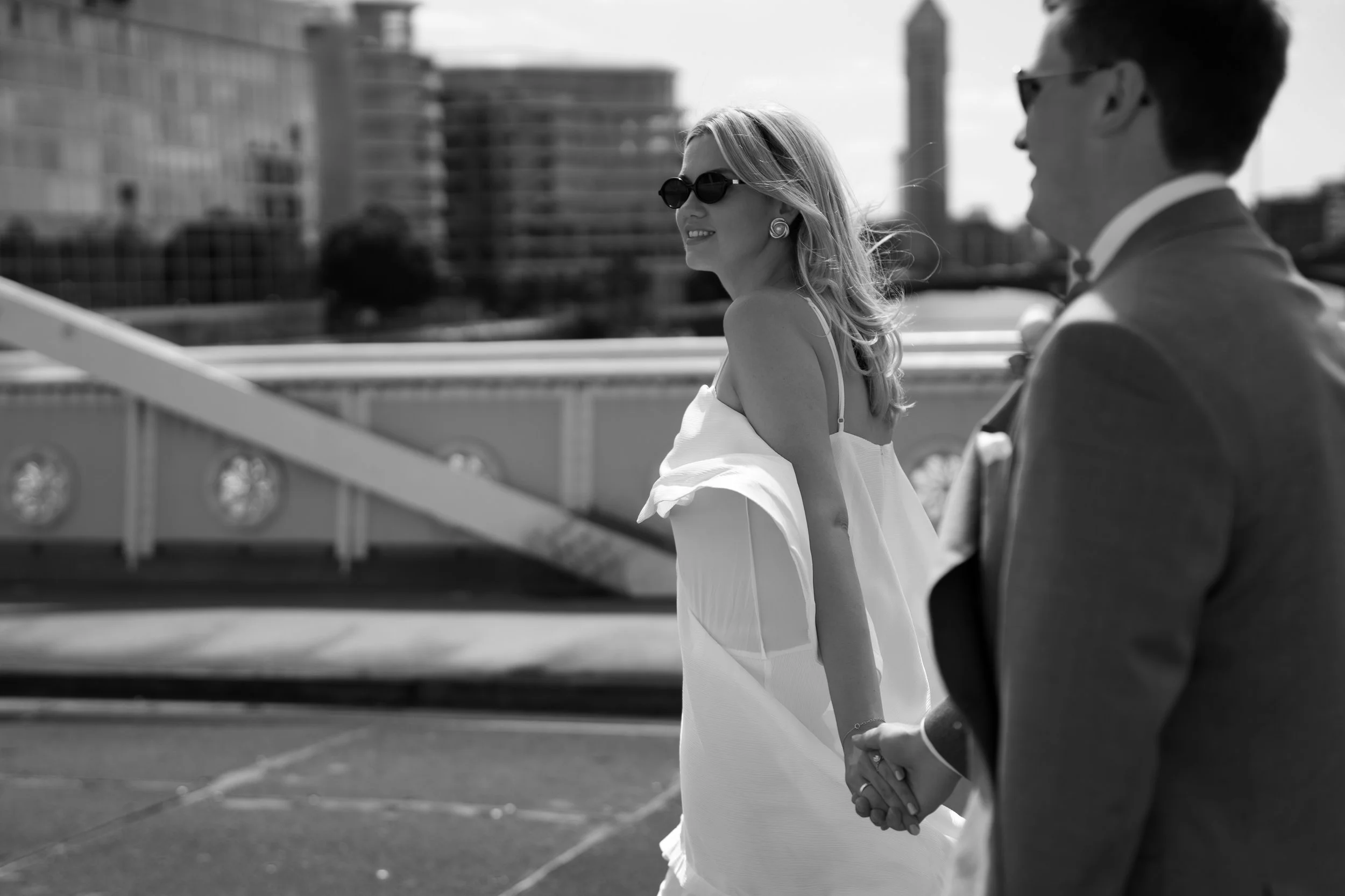 A woman in sunglasses and earrings holding hands with a man in a suit on a rooftop parking lot with buildings in the background.