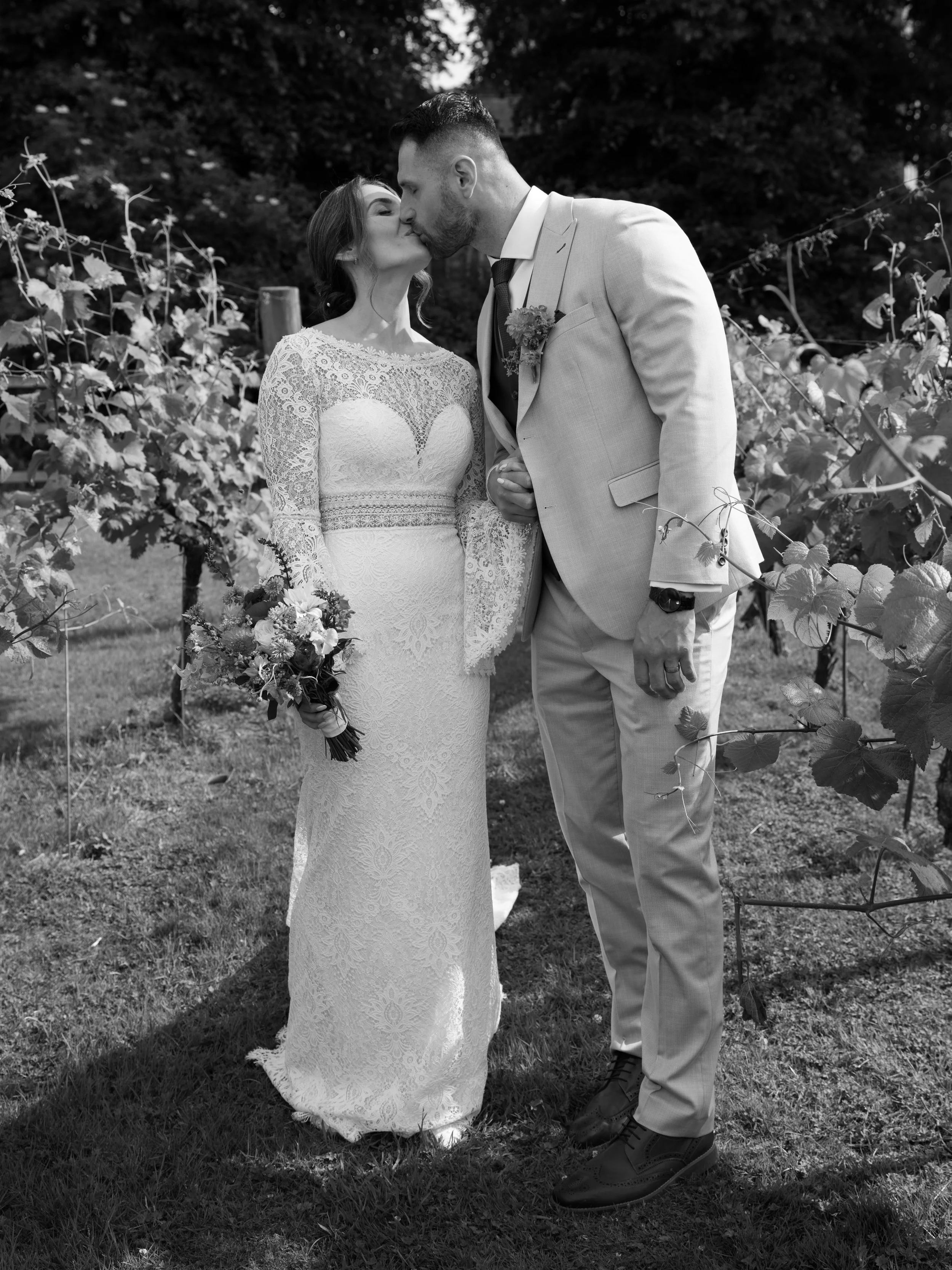 A black and white photograph of a bride and groom kissing outdoors in a vineyard. The bride wears a lace wedding dress and holds a bouquet, while the groom is dressed in a light-colored suit with a boutonniere.