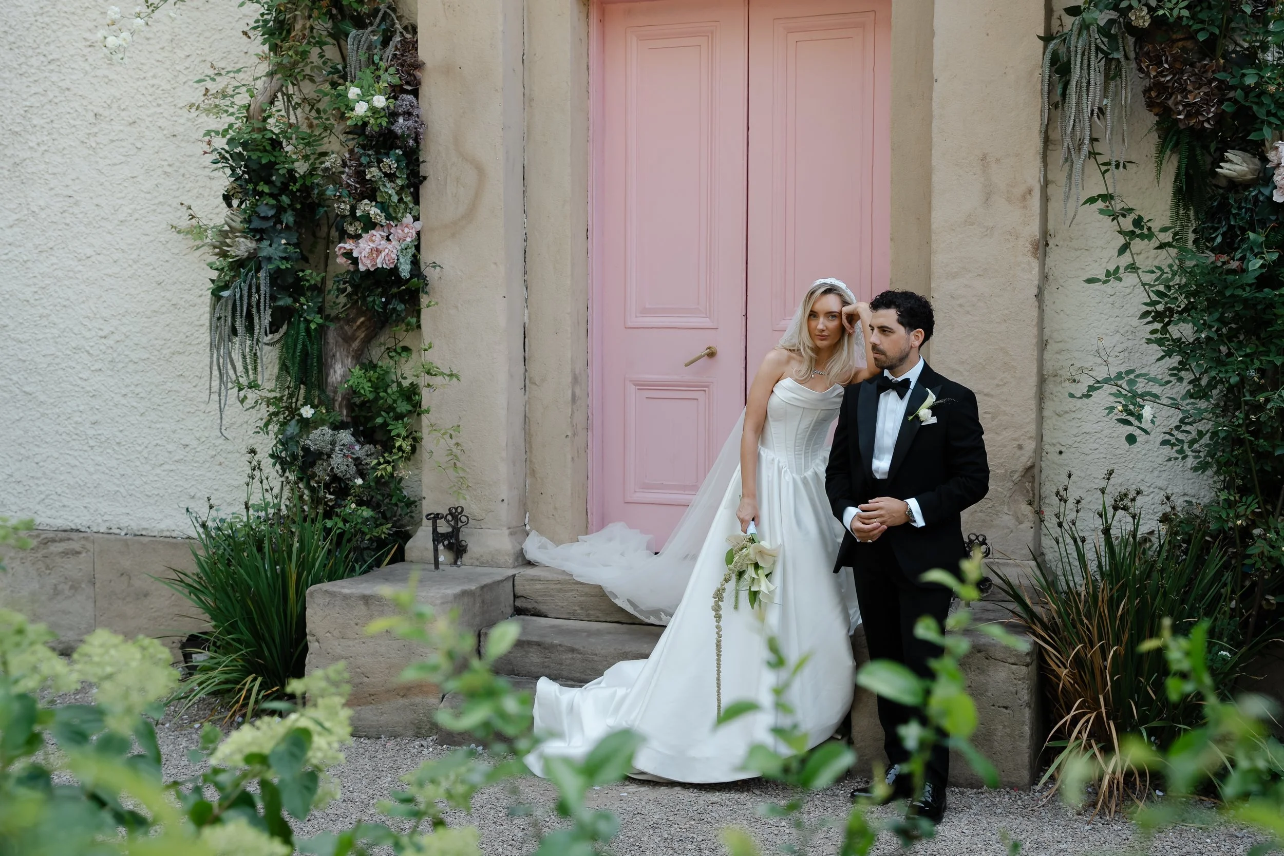 A bride and groom stand in front of a pink door, surrounded by greenery and floral arrangements, for a wedding photo.
