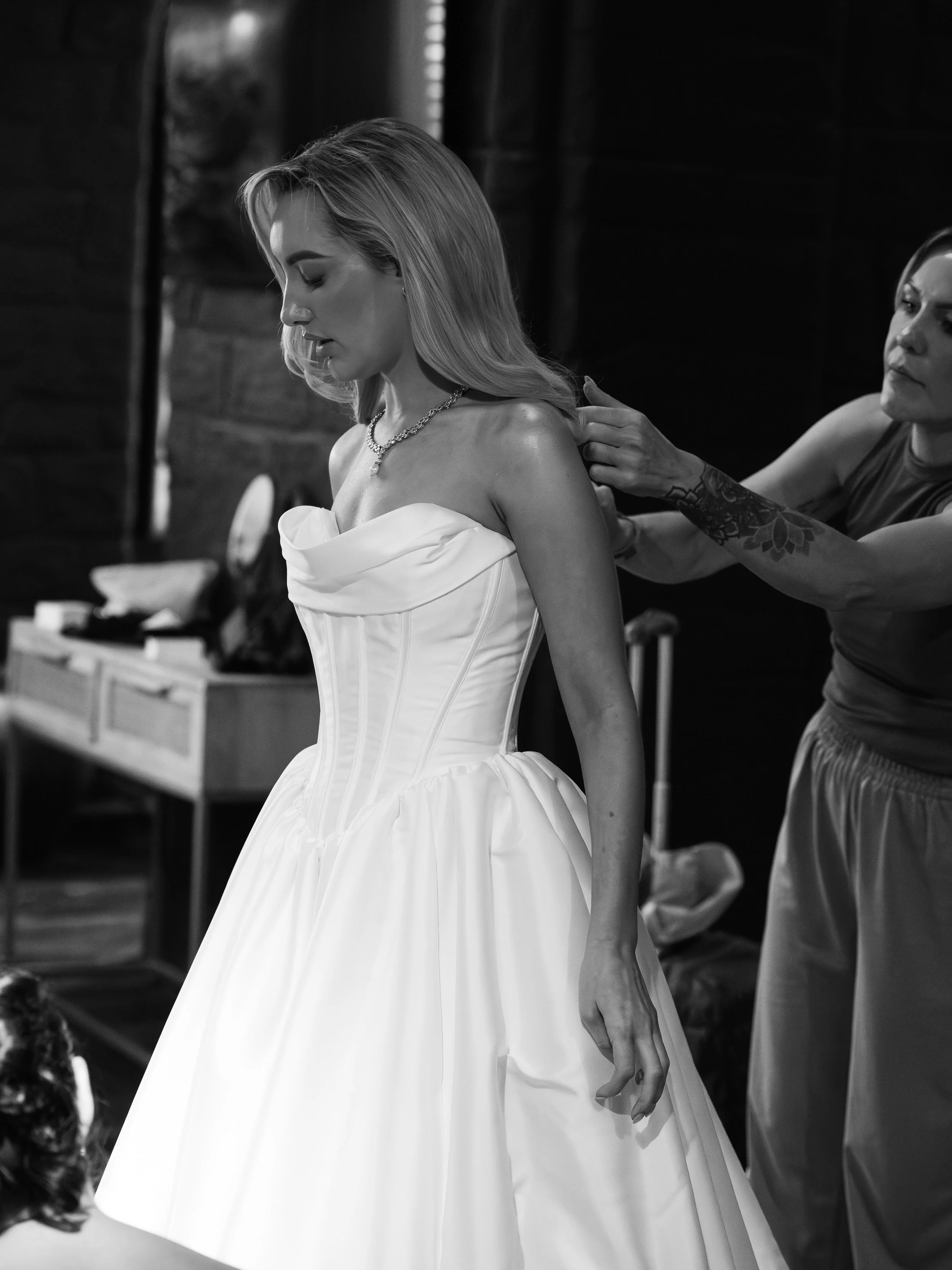 A woman in a white dress getting her back adjusted by a tattooed stylist in a backstage setting