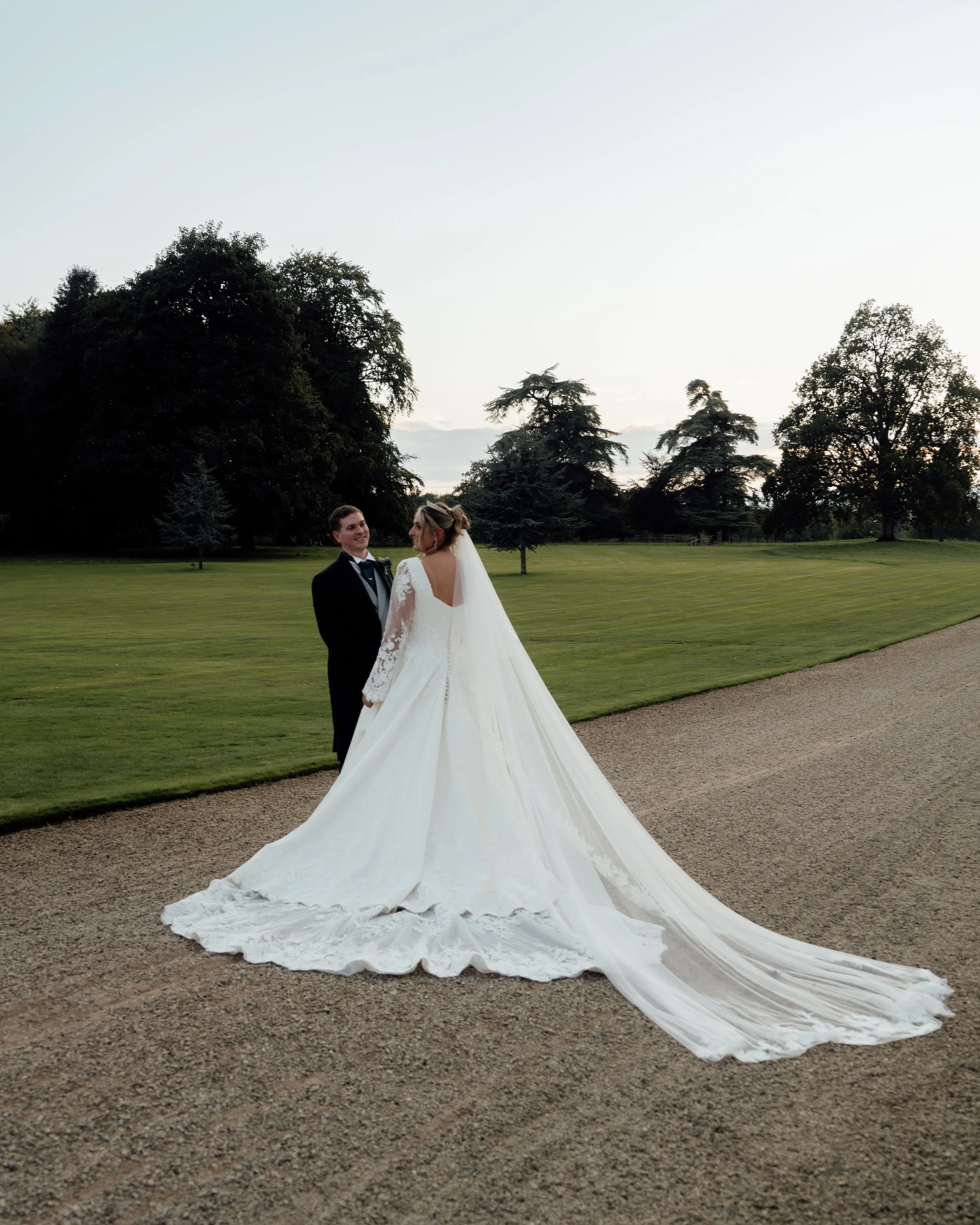 A bride and groom standing on a gravel pathway in a park with tall trees in the background during evening or late afternoon, celebrating their wedding.