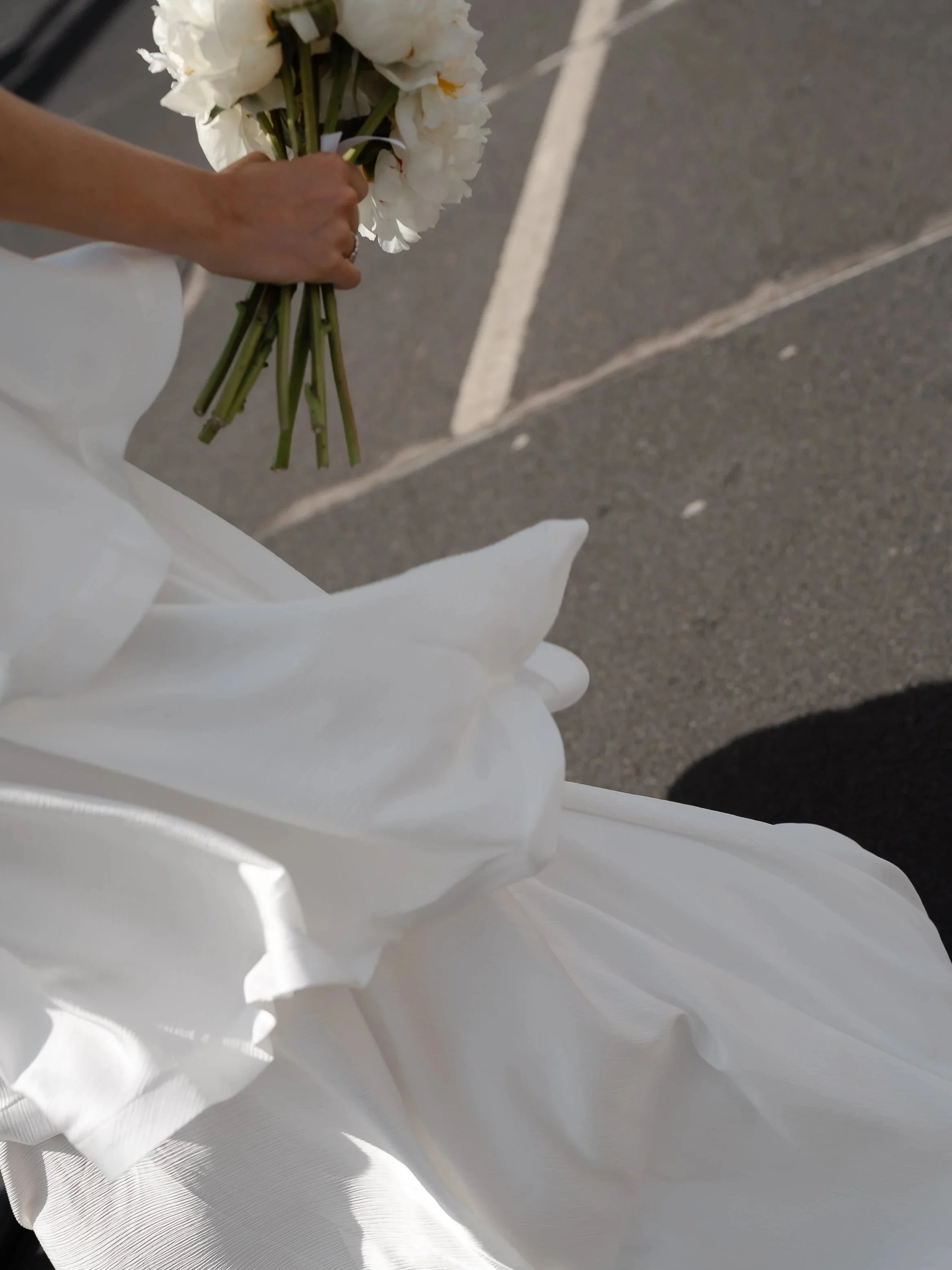 A person holding a bouquet of white flowers, possibly wedding or funeral flowers, with a white paper wrapping, captured on a paved surface.