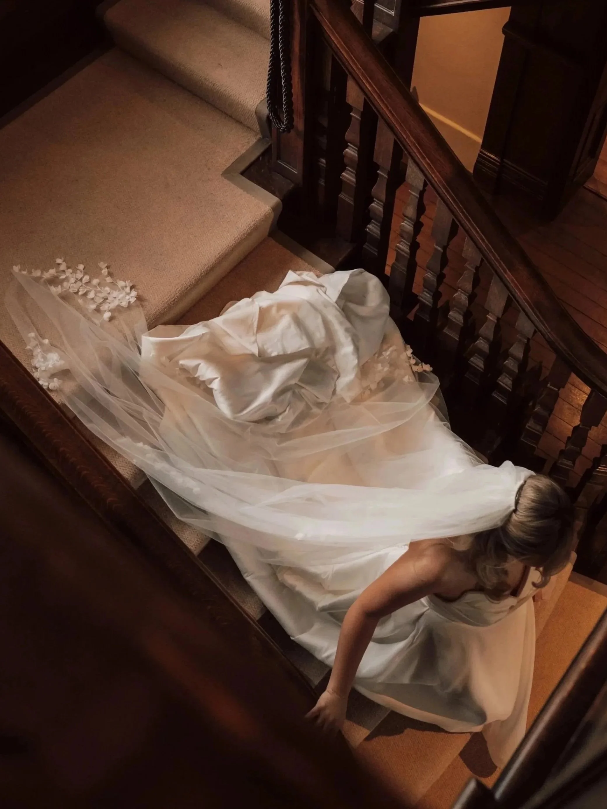 A woman in a wedding dress bending over on a staircase, holding her dress and touching the floor.