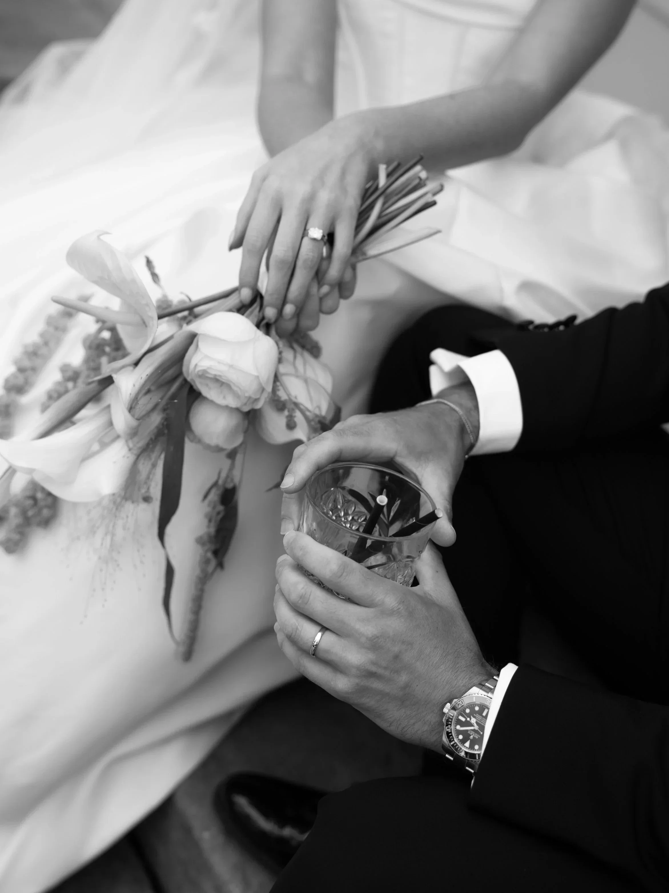 A person in formal attire with a wedding band on their finger is holding a flower bouquet with one hand and a glass of drink with a straw in the other hand. The person is wearing a wristwatch.