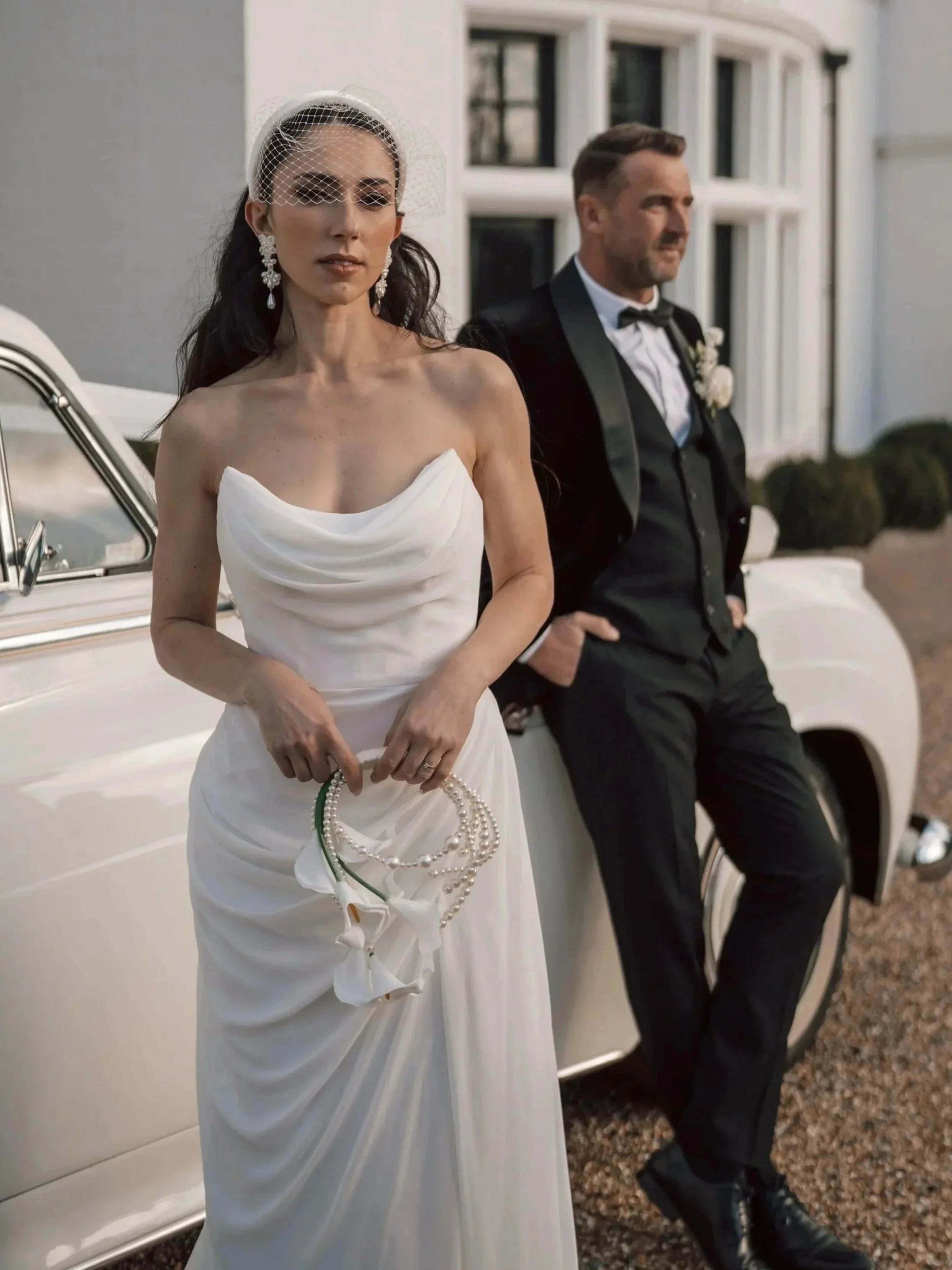 Bride in a white wedding gown and birdcage veil holding pearl necklaces and clutch, groom in a tuxedo with bowtie, standing next to a vintage white car outside a house with white windows.