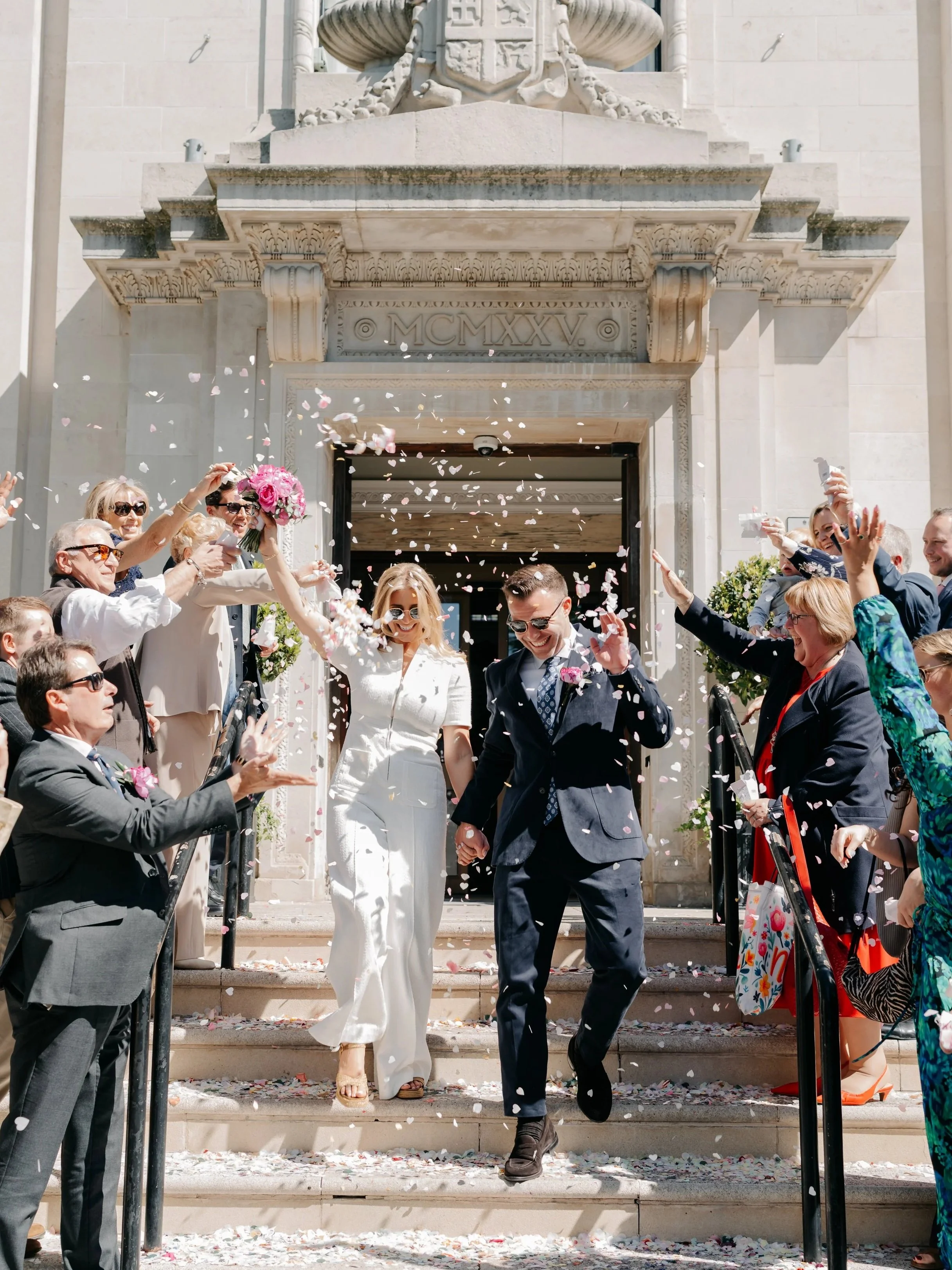 A wedding celebration with guests throwing flower petals as the bride and groom walk down the steps of a building with a decorative arch and winged emblem above the entrance.
