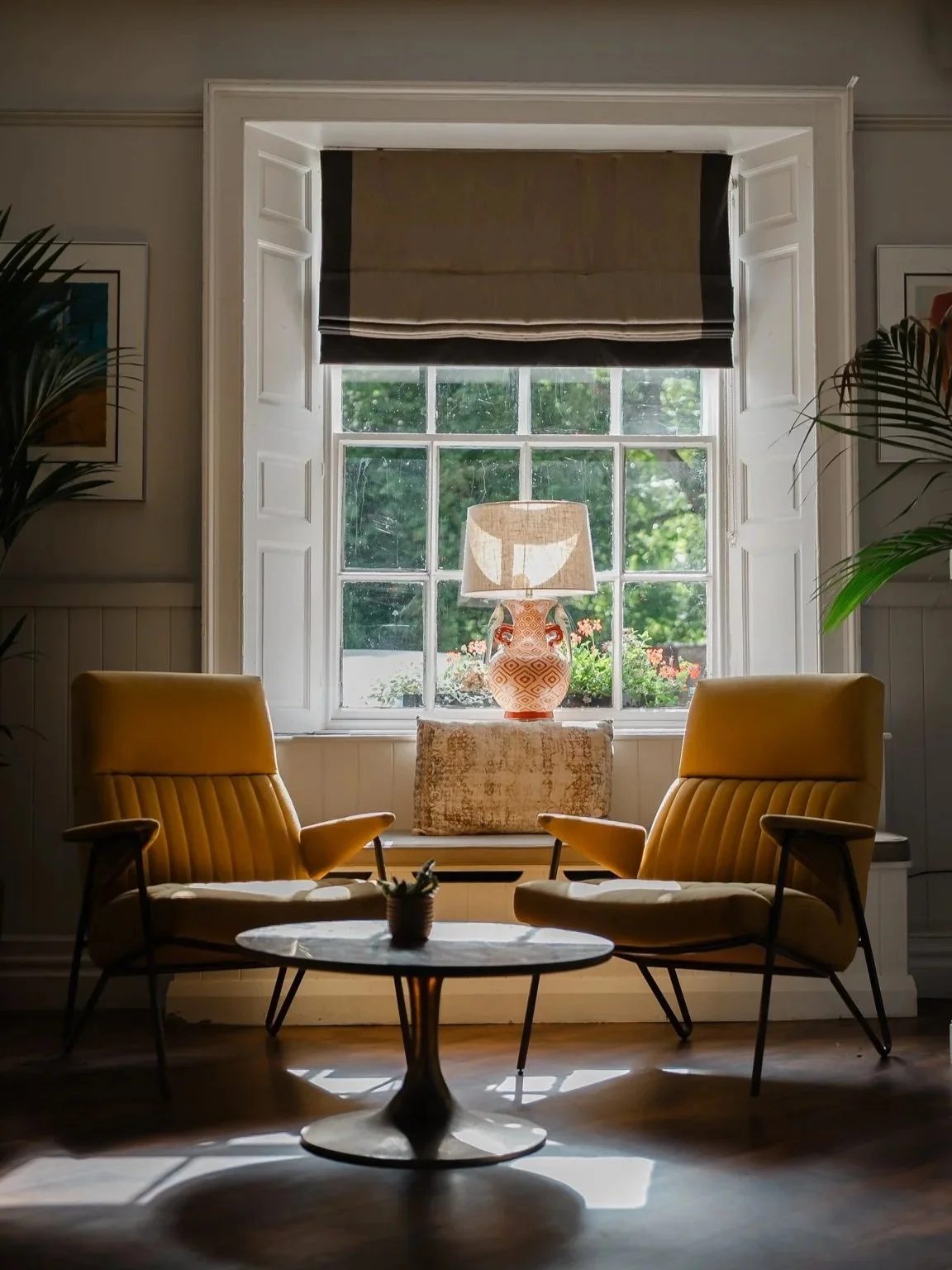 Cozy living room with two mustard yellow armchairs, a round marble-topped coffee table, a large window with a black roller shade, and a lampshade on a decorative vase on the windowsill, surrounded by green plants and framed artwork.