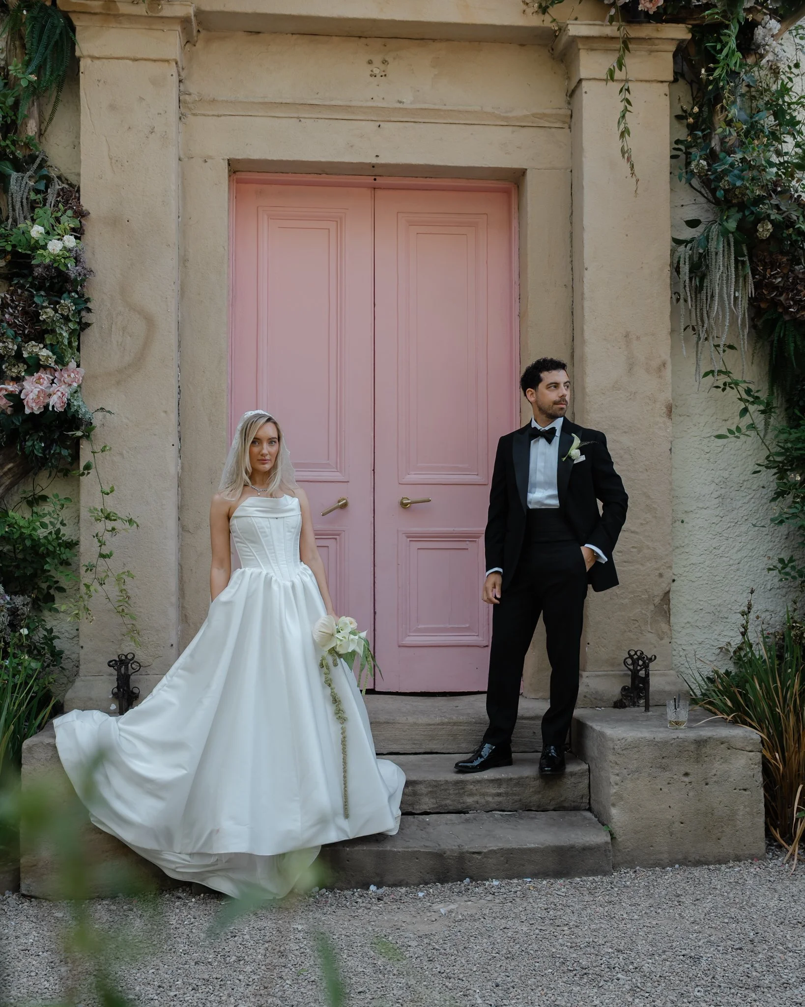 A bride in a white wedding gown with a strapless bodice and a veil, holding a bouquet of white flowers, standing on the steps in front of a pink door and an off-white wall. A groom in a black tuxedo with a bow tie and boutonniere is standing next to 