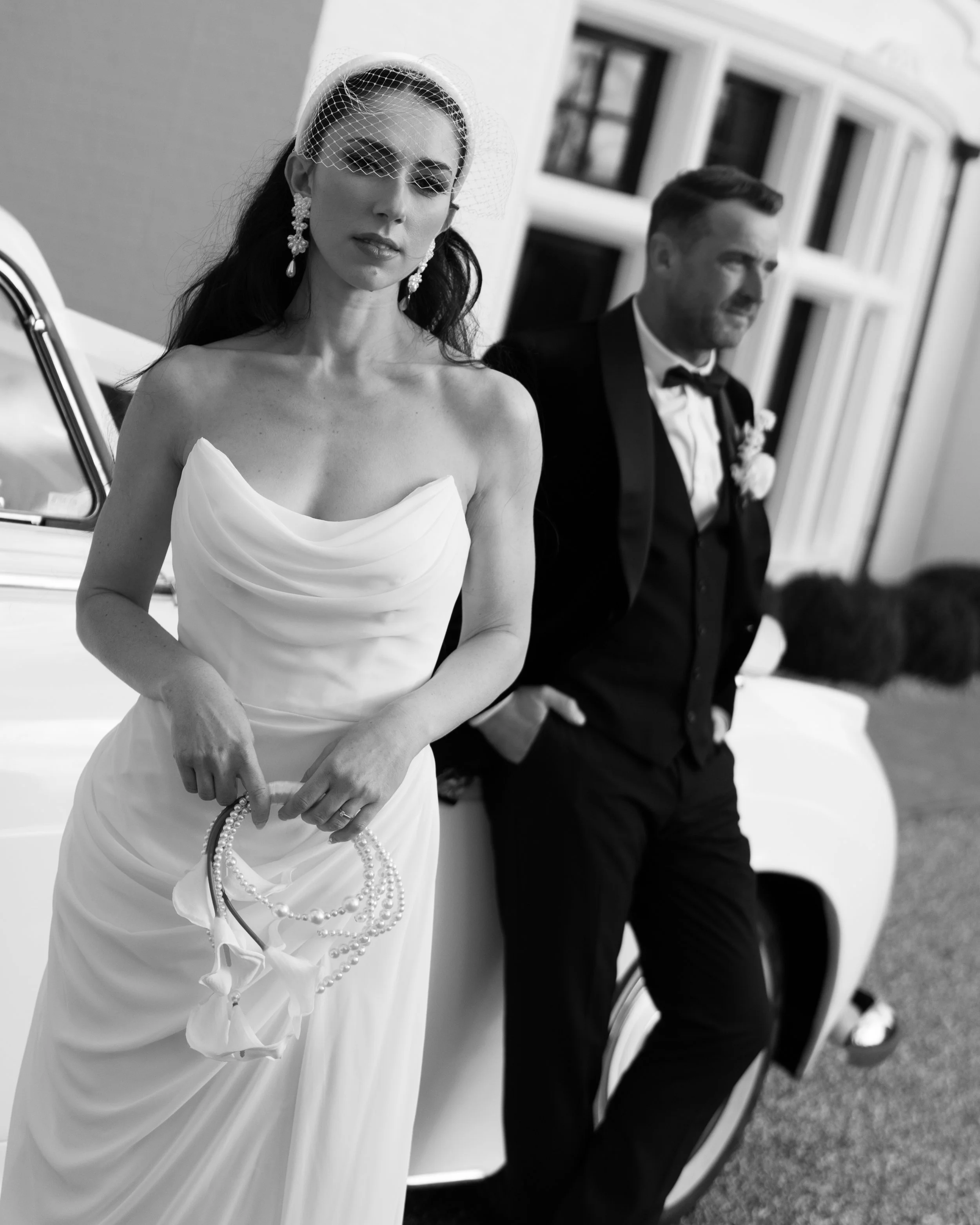 A woman in a strapless wedding dress holding a pearl necklace, standing next to a man in a tuxedo leaning against a vintage car, outside a house.