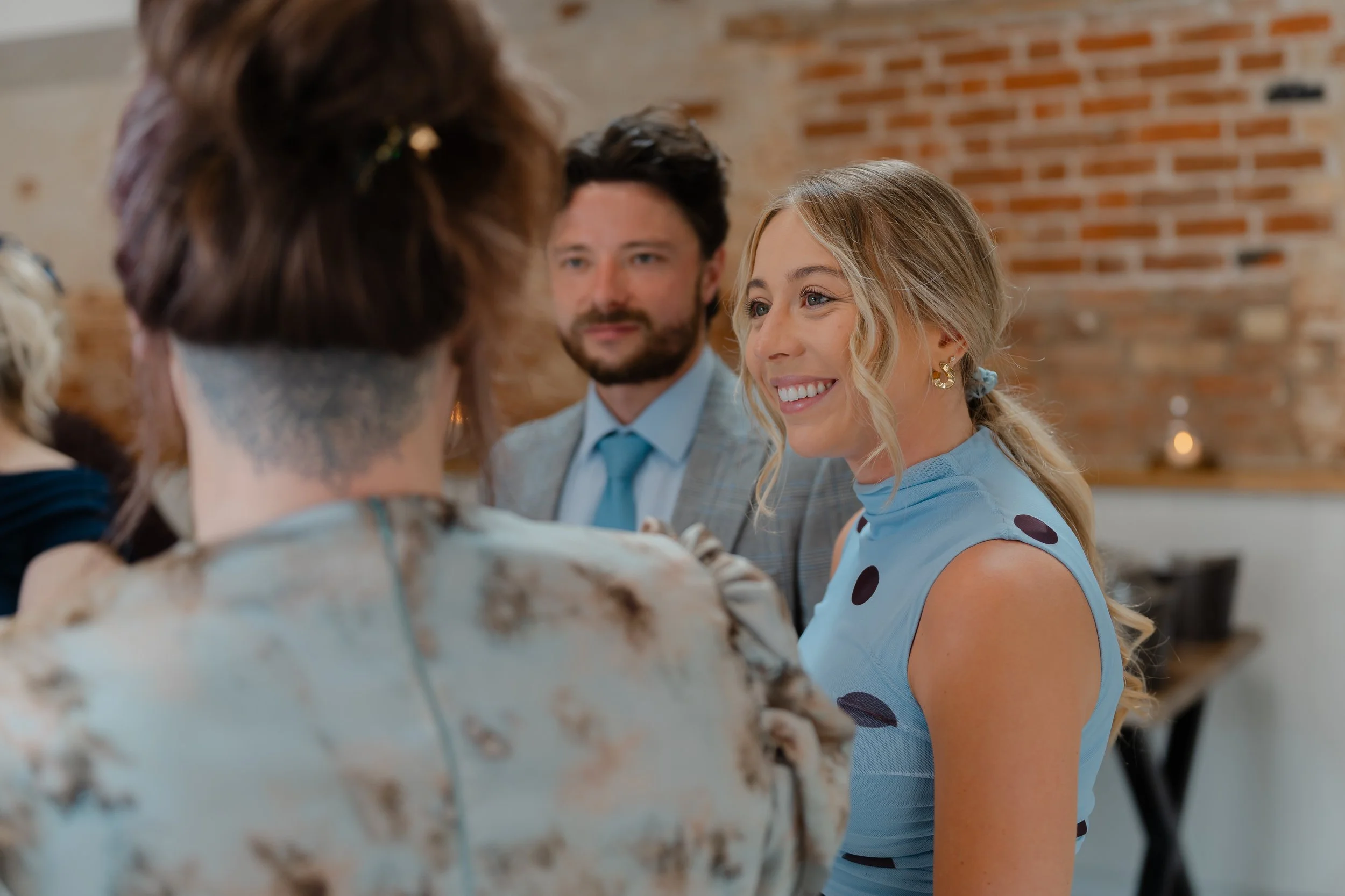 Four people at a social gathering, with two women and a man clearly smiling and engaged in conversation. One woman has blonde hair styled in loose waves and is wearing a light blue dress with dark polka dots. The man has dark hair and a beard, dresse