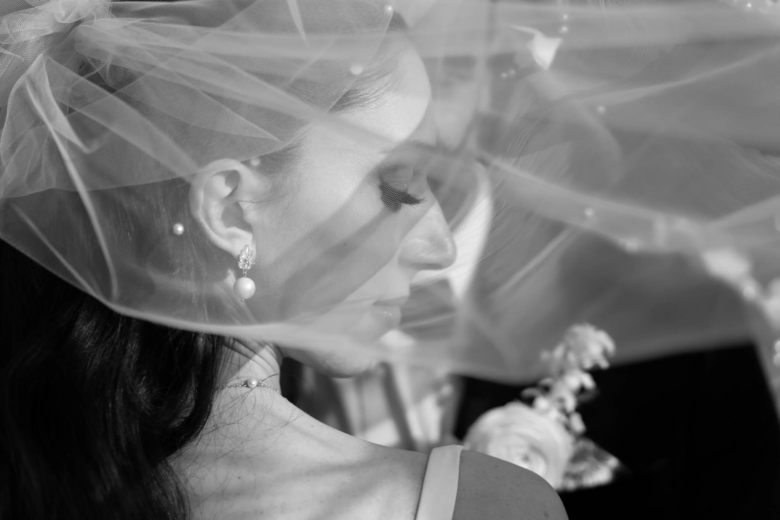 A bride in profile behind a veil, with her eyes closed and wearing pearl jewelry.