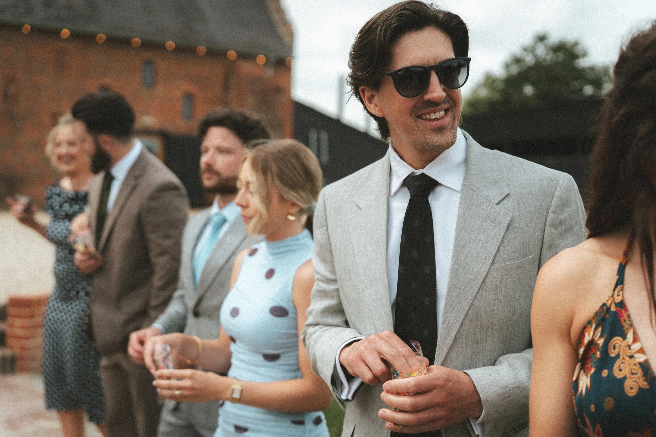 A group of people dressed in formal and semi-formal attire socializing at an outdoor event, with some holding drinks, in front of a rustic brick building with string lights.