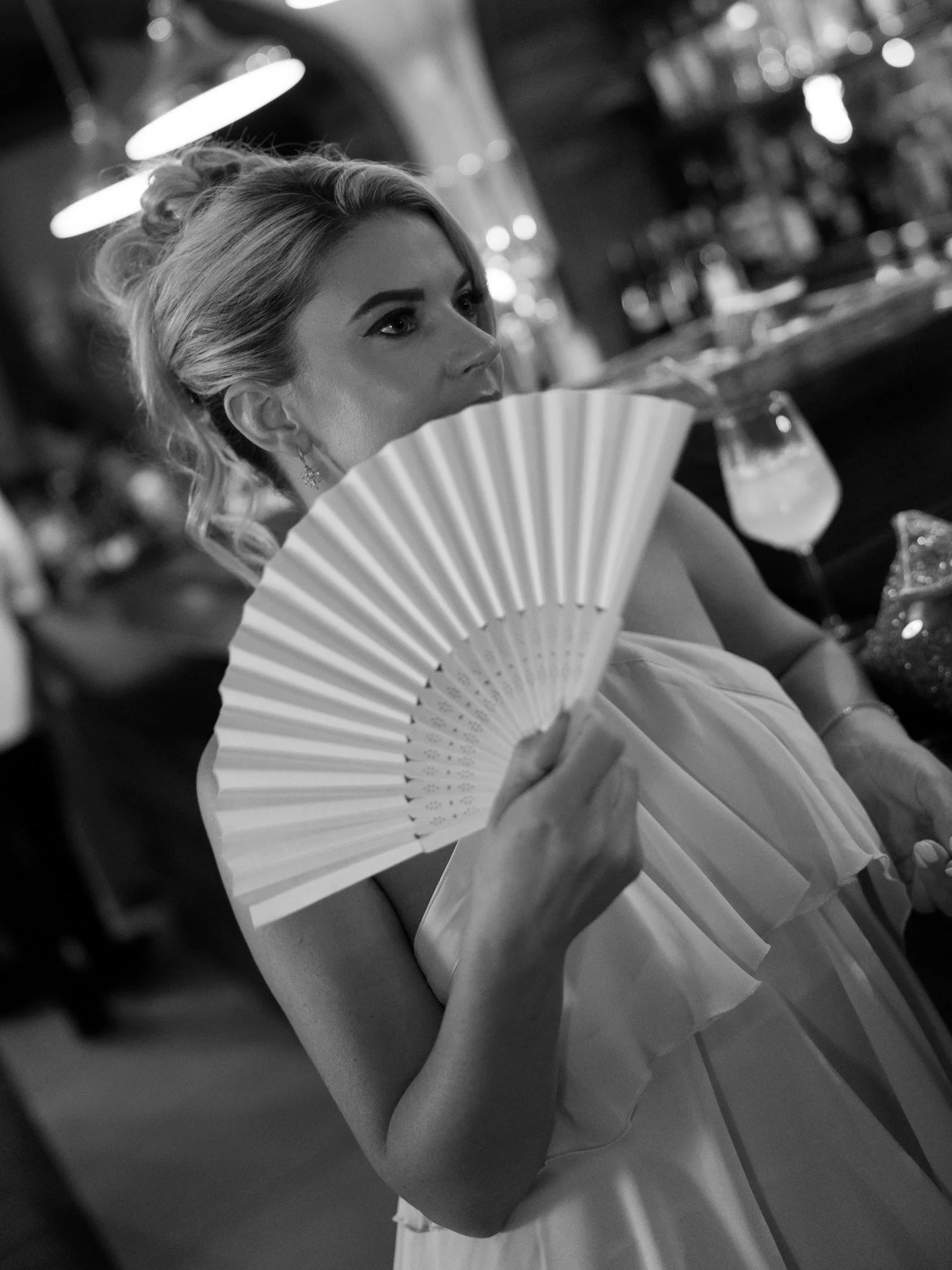 A black and white photo of a woman holding a traditional folding fan close to her face, with a blurred background of a bar or restaurant.