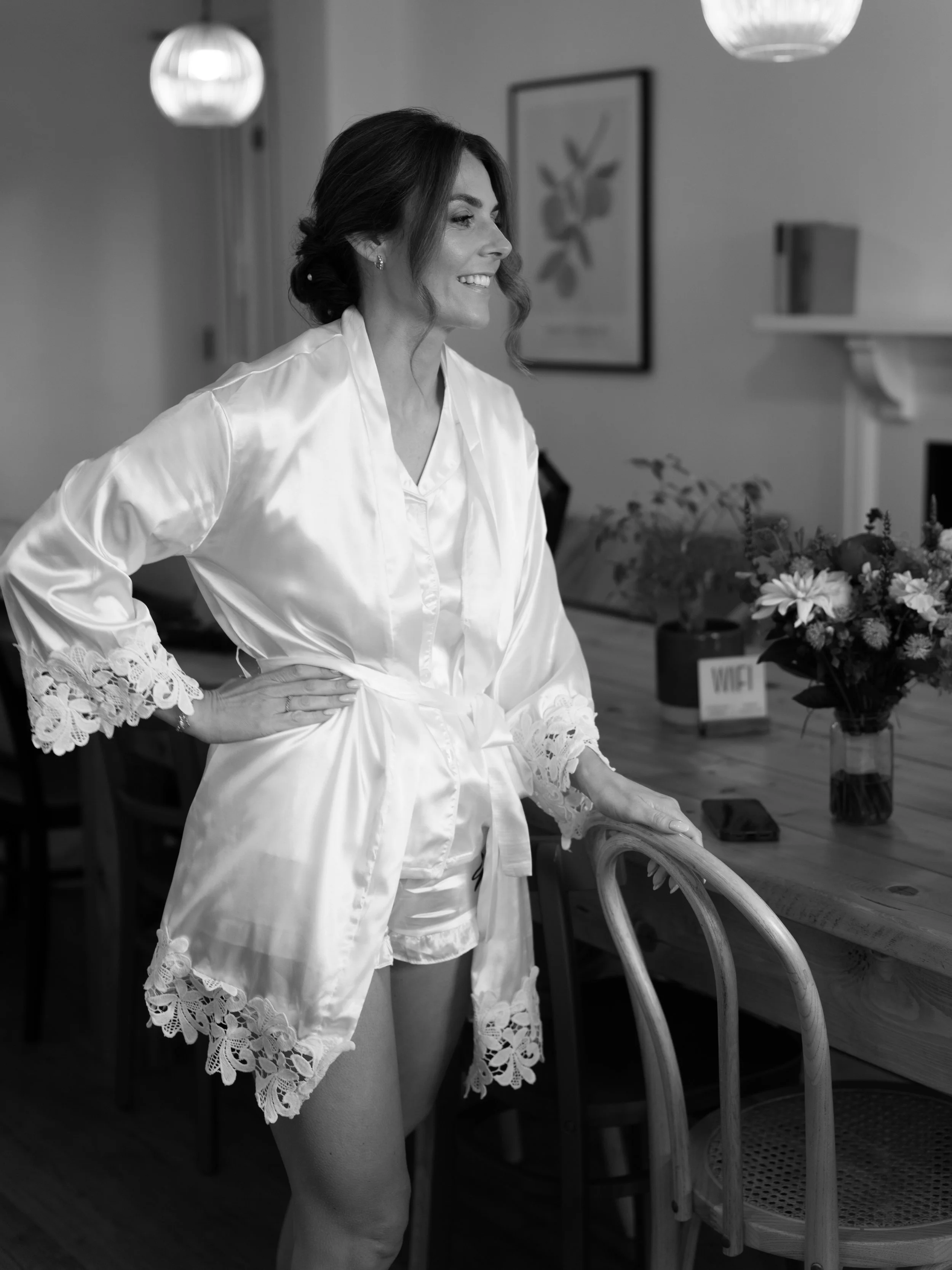 A bride in satin and lace lingerie stands in a room, smiling, with her hands on her hips, next to a wooden chair and a table with flowers in the Copdock Hall House