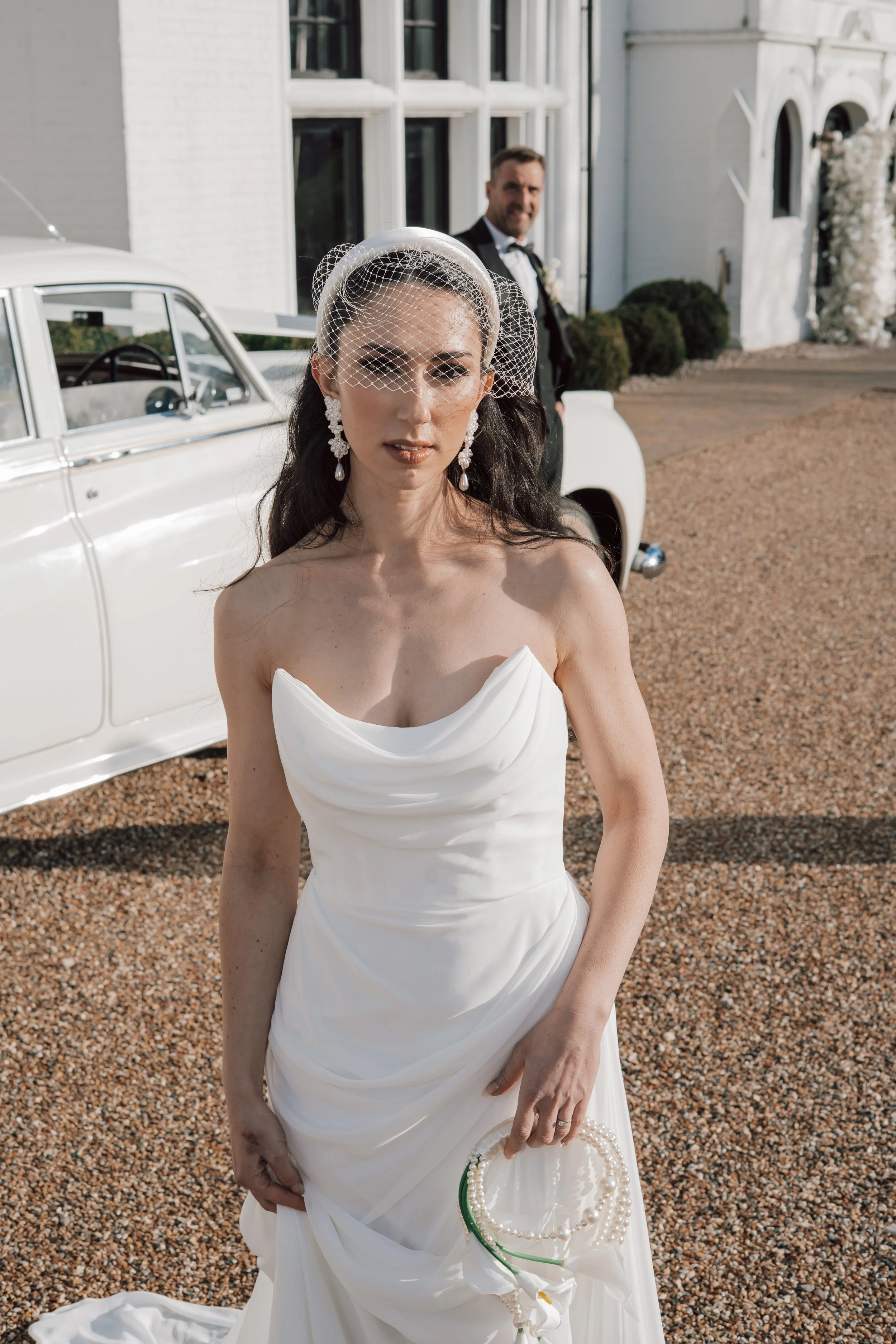 A bride in a strapless white wedding dress holding a pearl necklace, standing outdoors in front of a vintage white car, with a groom in a tuxedo in the background.