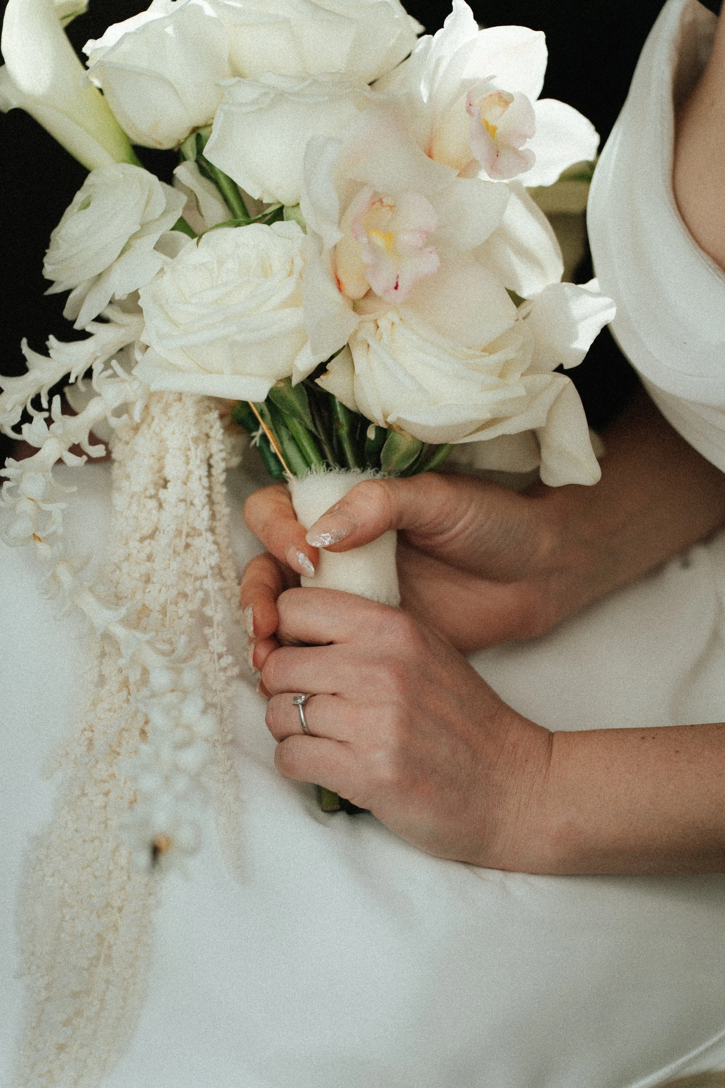 Close-up of a woman's hands holding a bouquet of white roses, orchids, and other flowers, with a wedding ring visible on her finger.