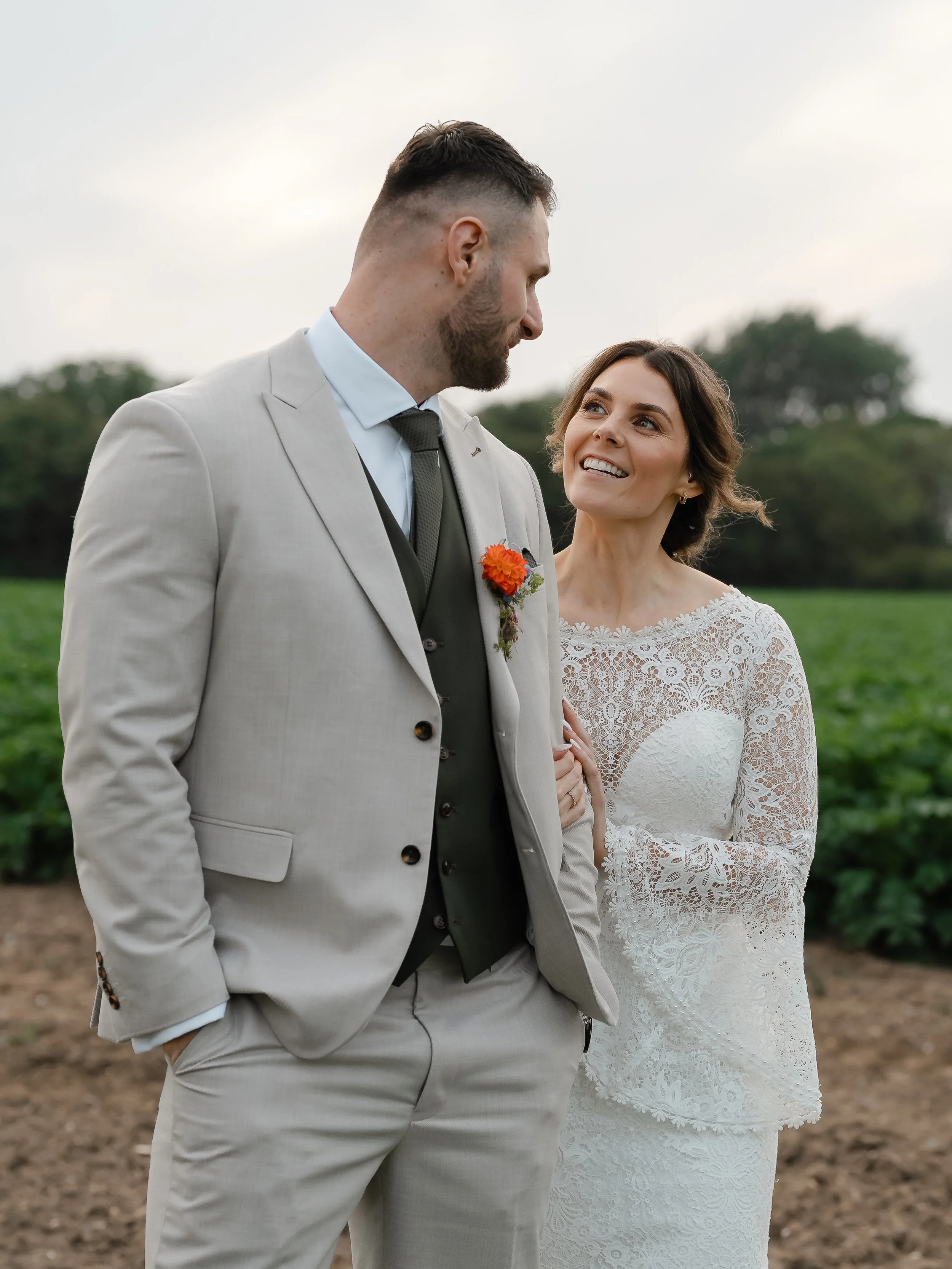 A bride and groom standing outdoors in a garden, smiling and looking at each other. The groom is wearing a light gray suit with a boutonnière, and the bride is in a white lace wedding dress with short sleeves.