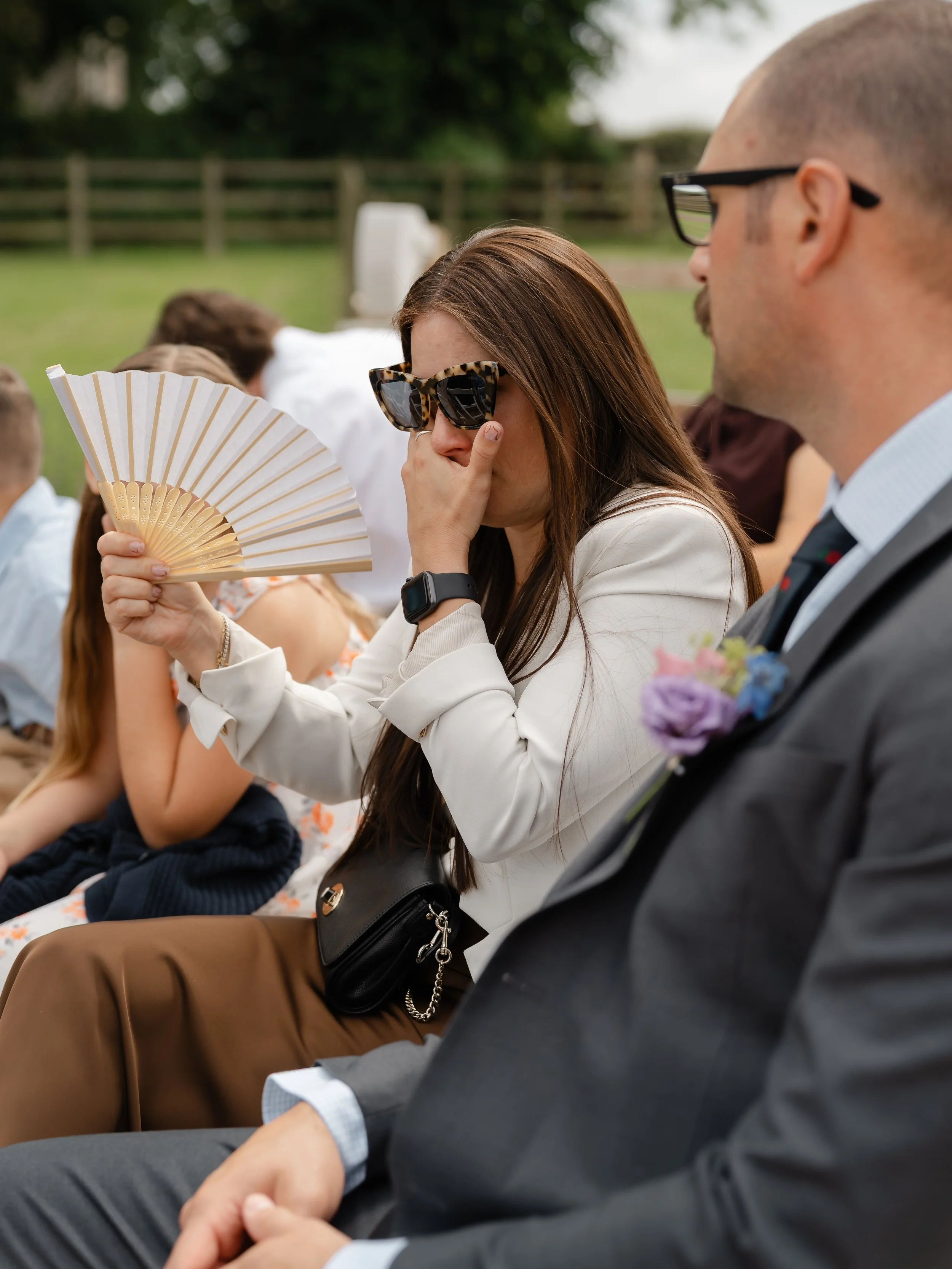 People sitting outdoors, some wearing sunglasses, with a wooden fence and greenery in the background.